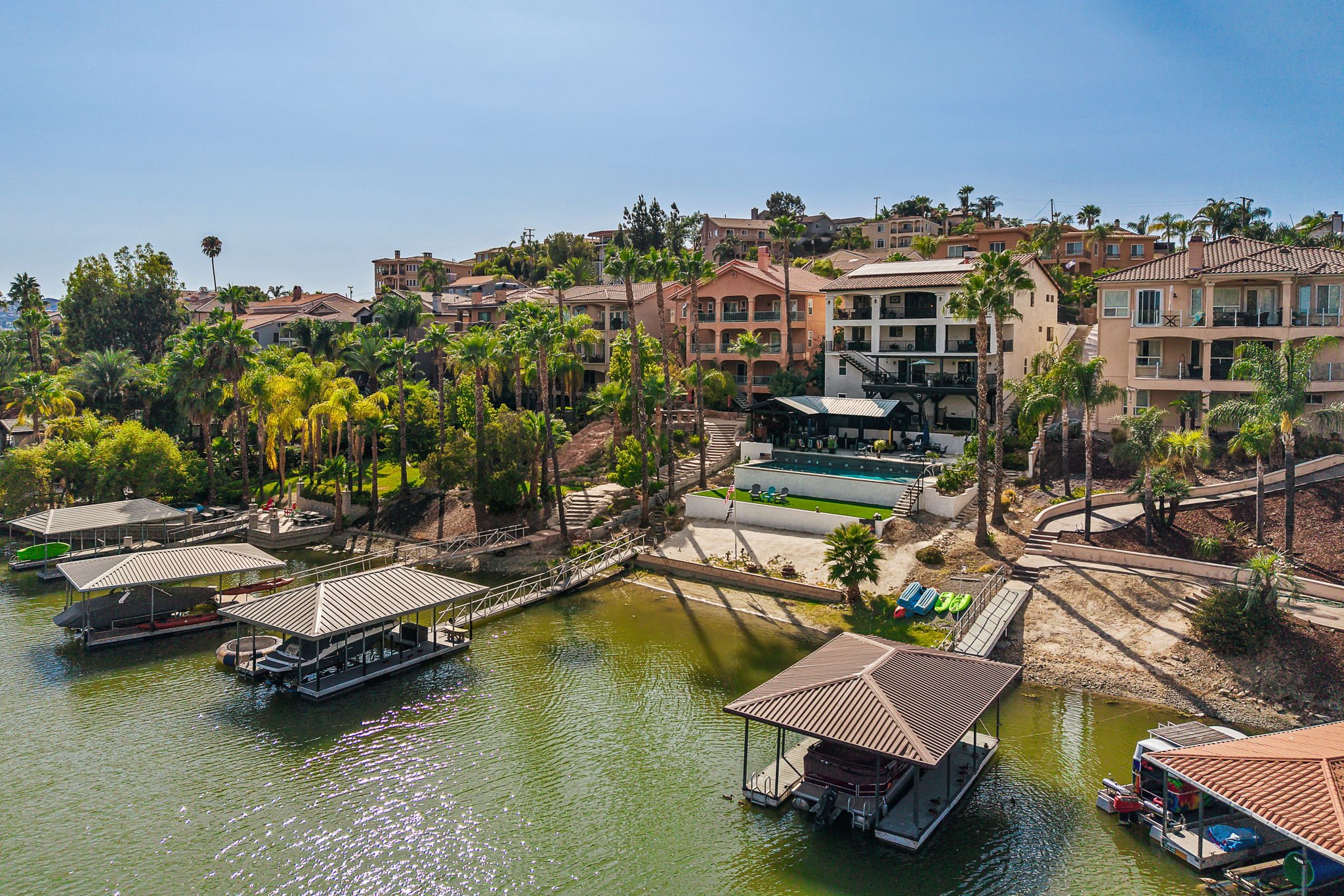 A view of a residential neighborhood with multiple multi-story houses, lush green palm trees, and a waterway with boat docks and covered boat slips.