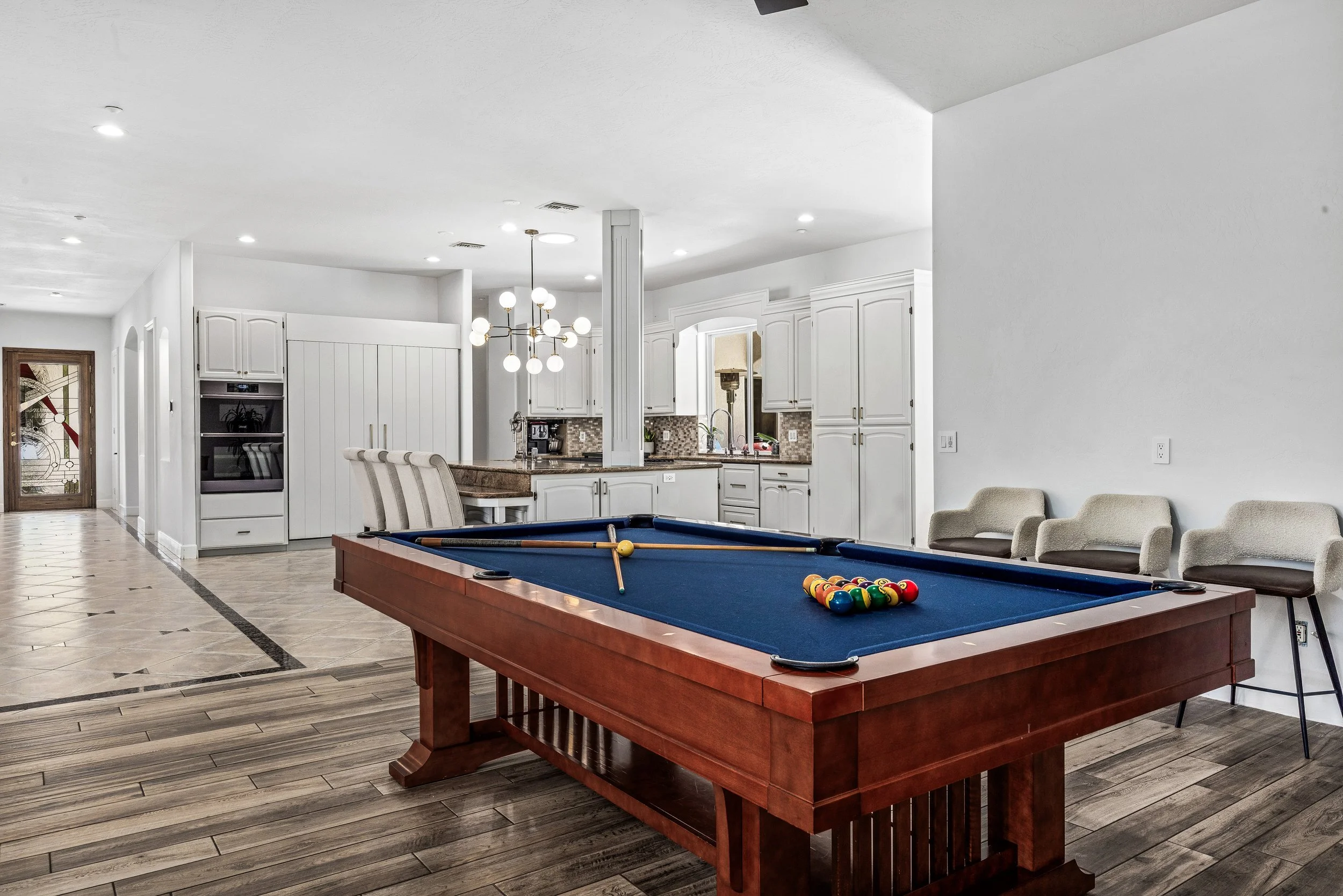 A game room with a blue pool table and a set of billiard balls, chairs along the wall, and a kitchen area in the background with white cabinets and a chandelier.