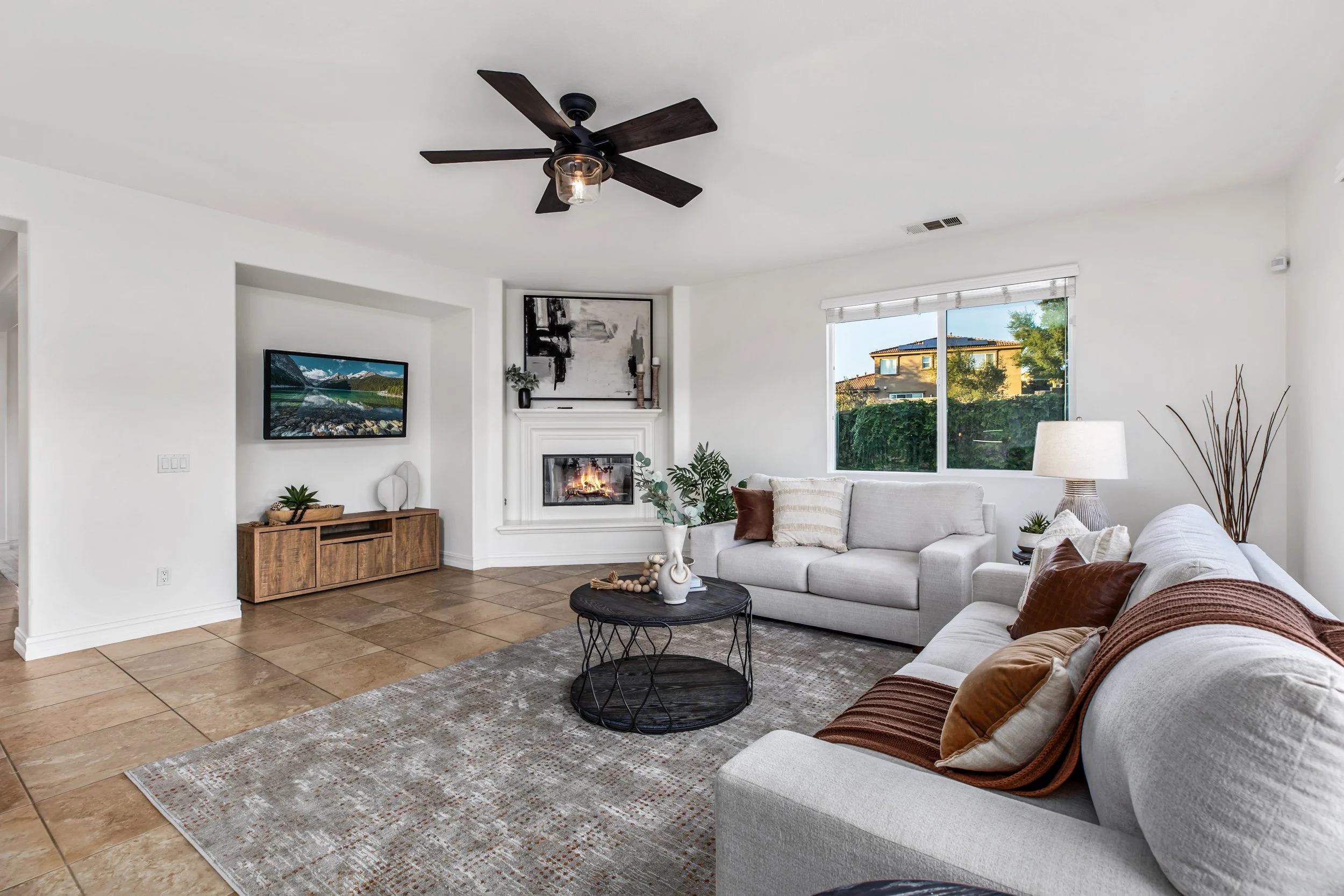 Living room with white walls, tiled floor, ceiling fan, fireplace, window, sofas, coffee table, decor items, and artwork.