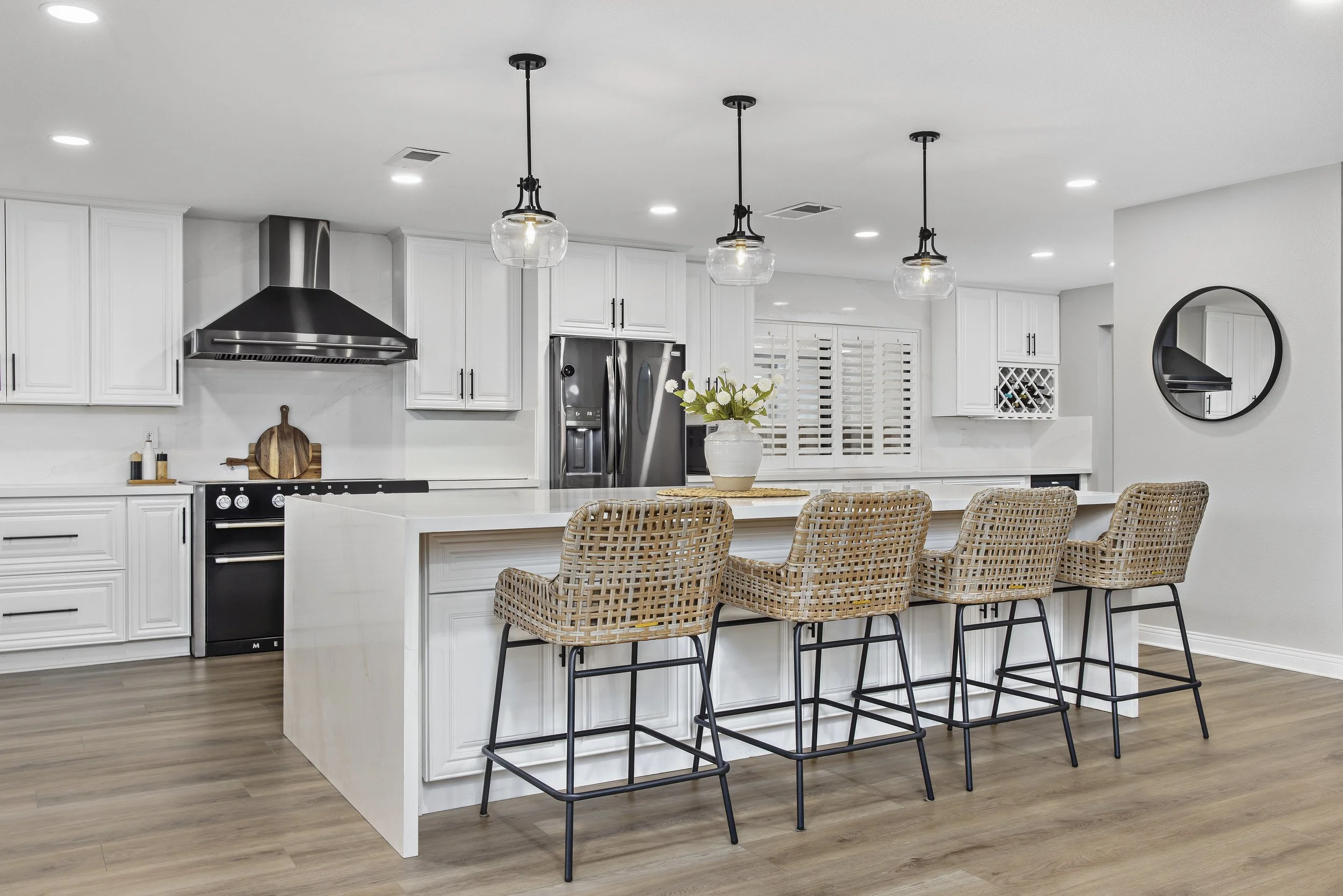Modern kitchen with white cabinetry, stainless steel appliances, and a kitchen island with woven barstools. Three pendant lights hang above the island, and there is a vase with flowers on the counter. The kitchen has hardwood floors and a large window with white shutters.