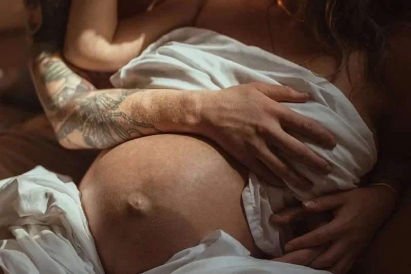 A woman with a tattooed arm and pregnant belly lying on a bed, holding her belly.