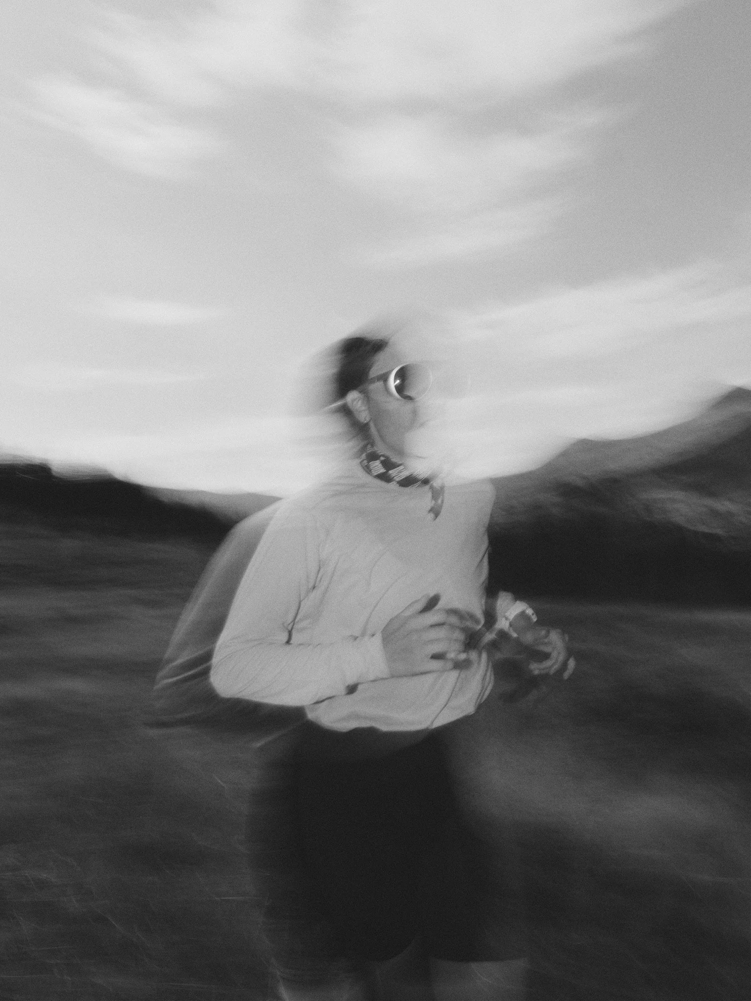 A blurred black and white photo of a person running outdoors, wearing sunglasses, a long sleeve shirt, and holding a camera or binoculars, with mountains and sky in the background.