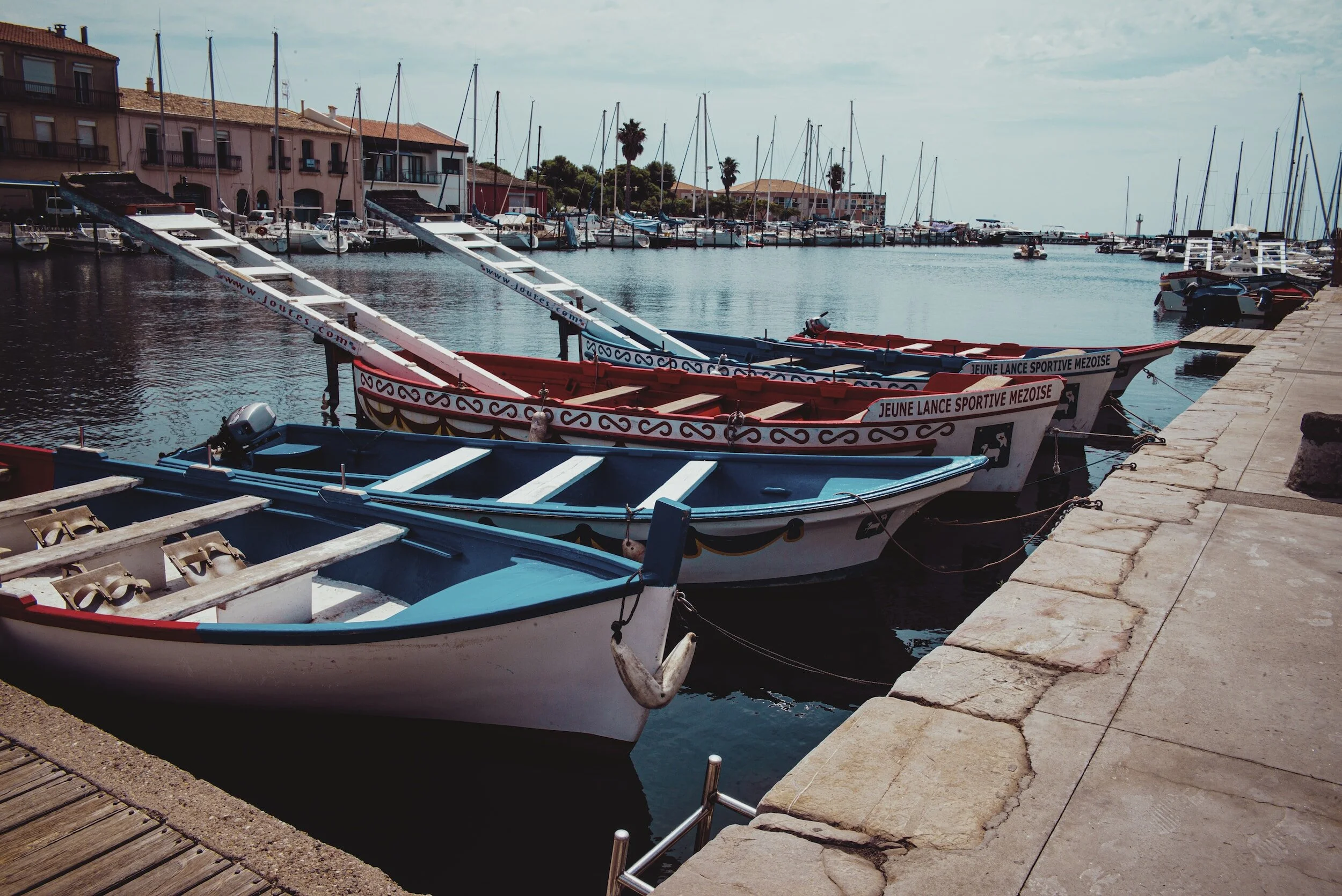 Bateaux traditionnels colorés amarrés dans un port, avec des bâtiments en arrière-plan et des mâts de voiliers visibles.