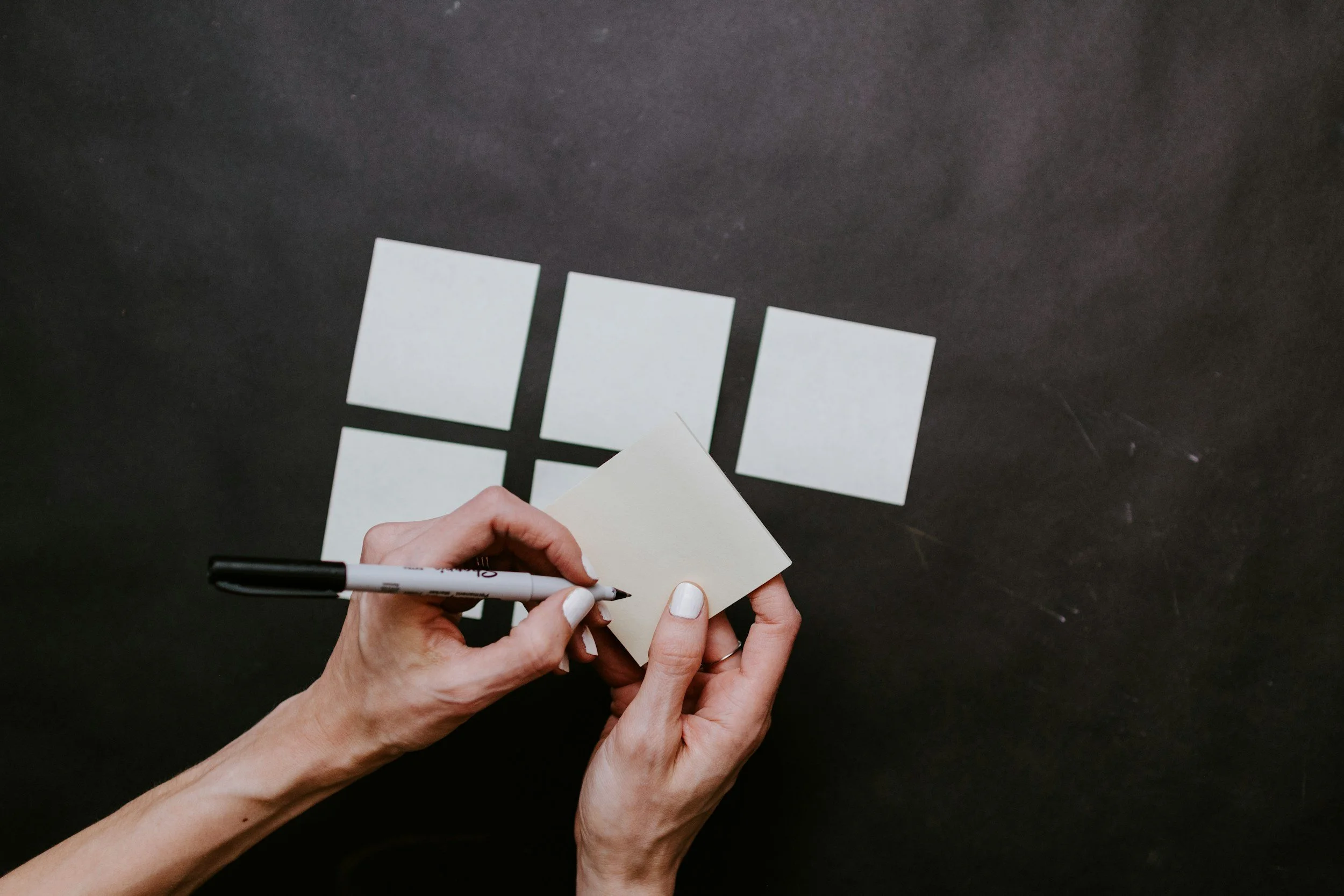 Person's hands holding a black dry-erase marker and a small sticky note, arranging sticky notes on a black table to form a 2x3 grid that resembles a window.