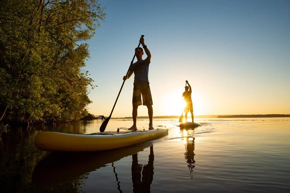 Twee mensen paddleboarden op een meer bij zonsondergang, met bomen langs de oever en een heldere lucht.