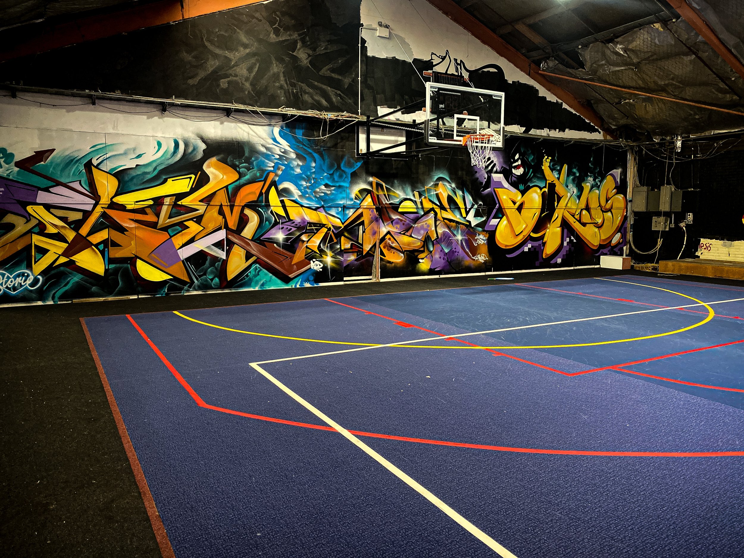 Indoor basketball court with colorful graffiti artwork on the wall and a basketball hoop.