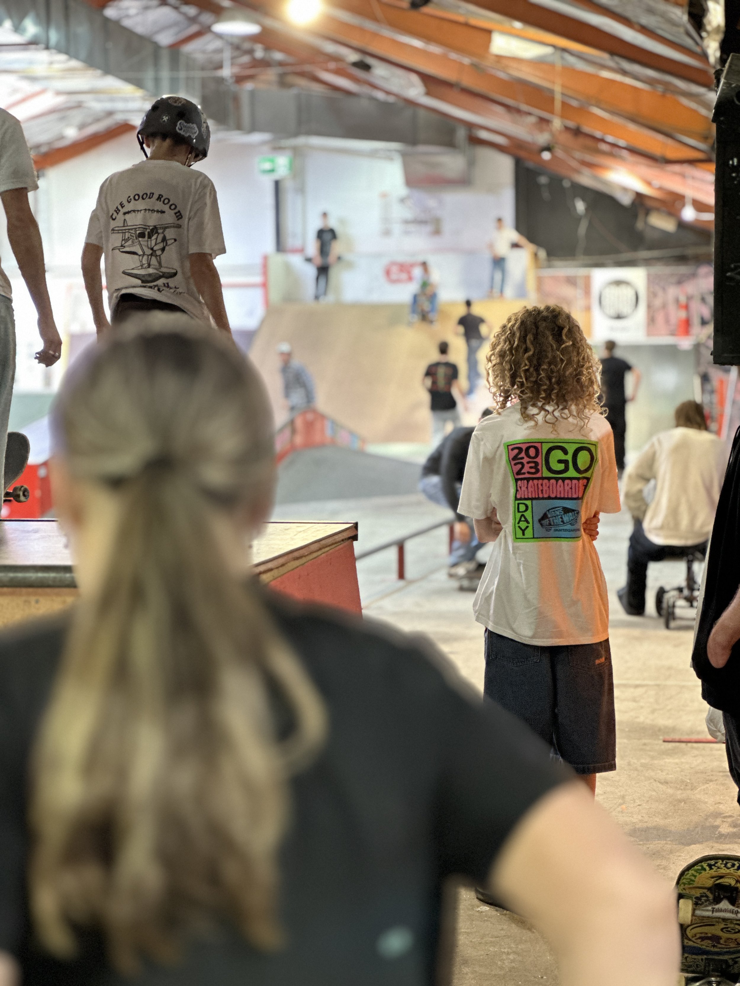 People inside a skate park with ramps, observing and preparing to skateboard. The focus is on individuals wearing casual clothing, including a person with curly hair wearing a shirt with colorful text. The skate park has a high ceiling and bright lighting.