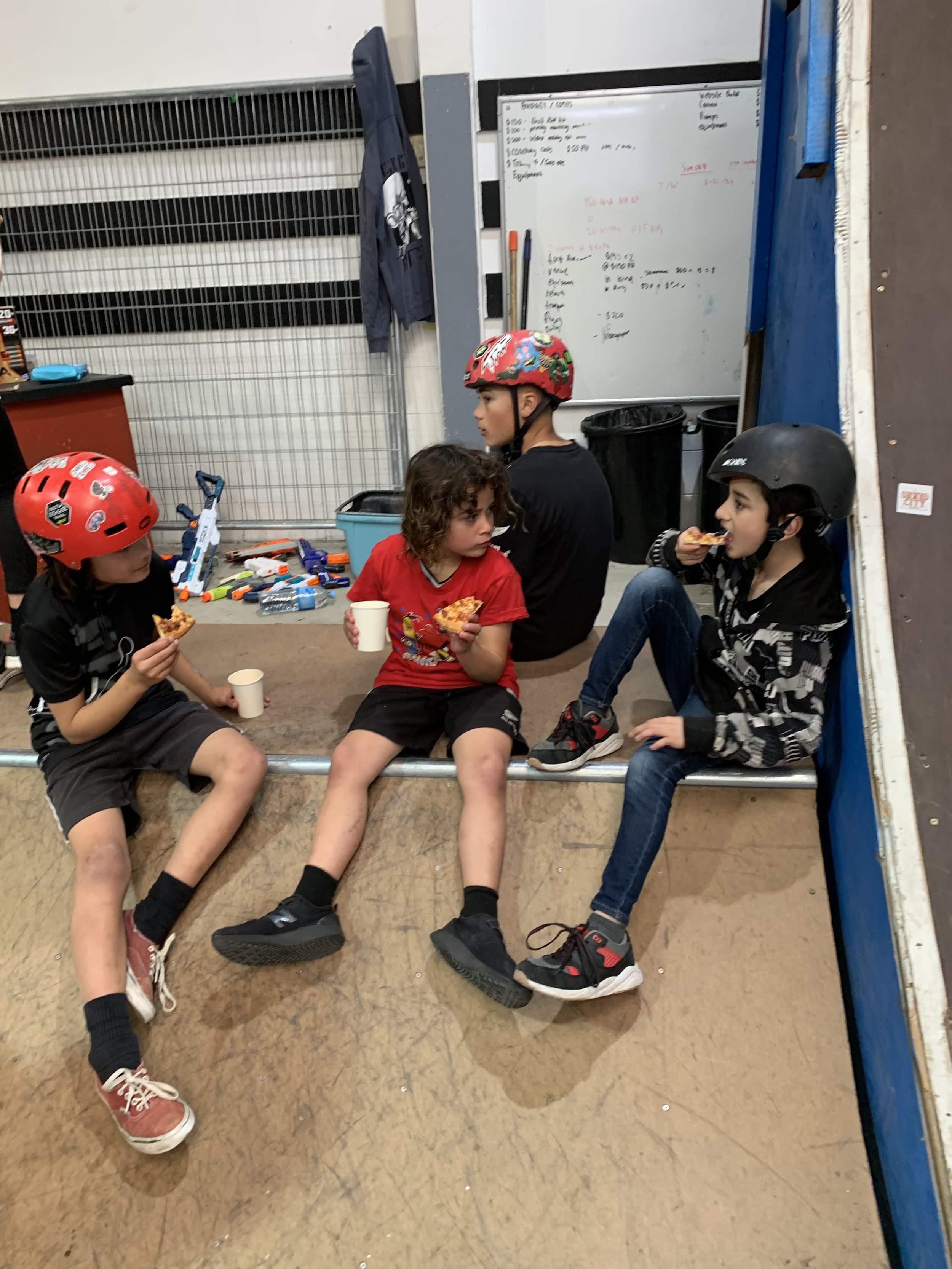 Kids wearing helmets sitting on a skateboard ramp eating pizza.