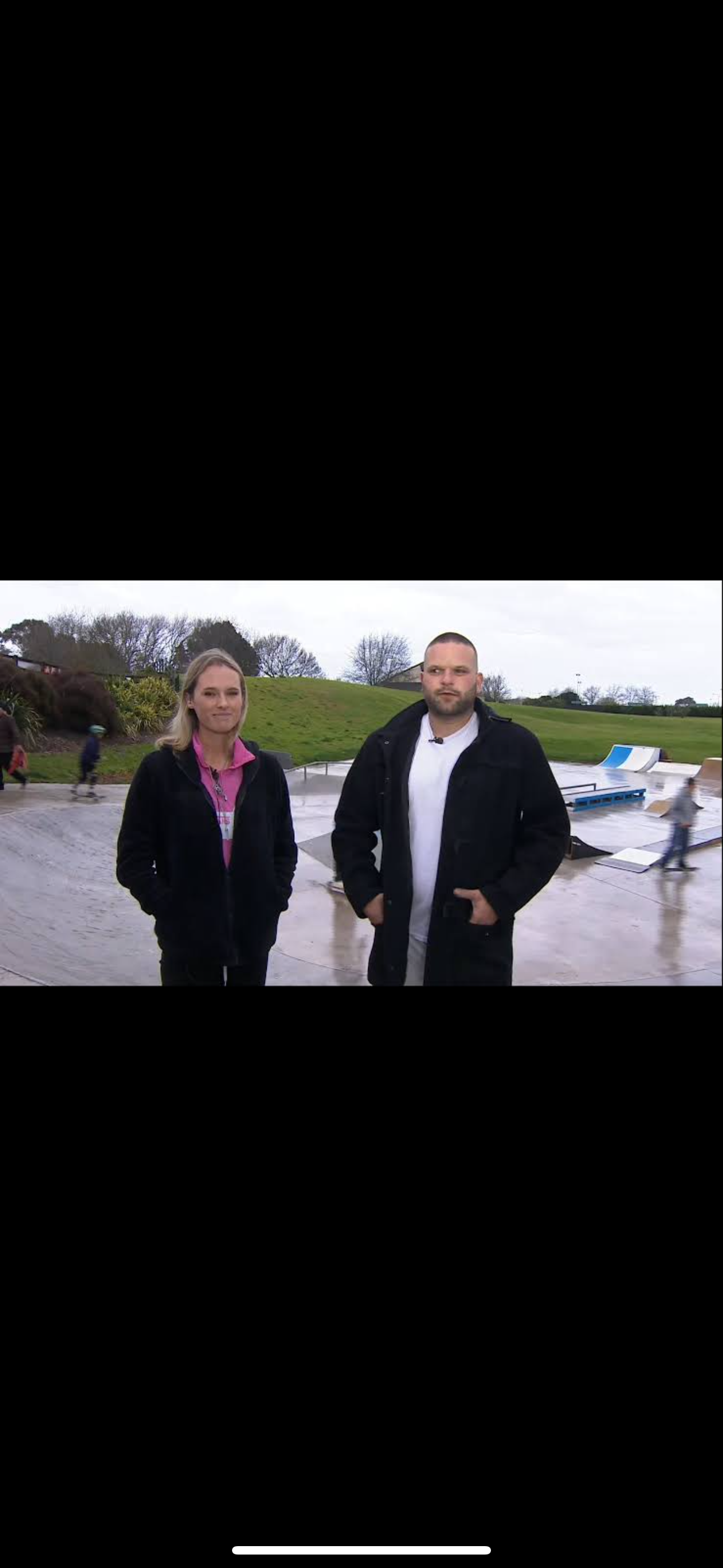 Two people standing in a skate park with ramps and greenery in the background.