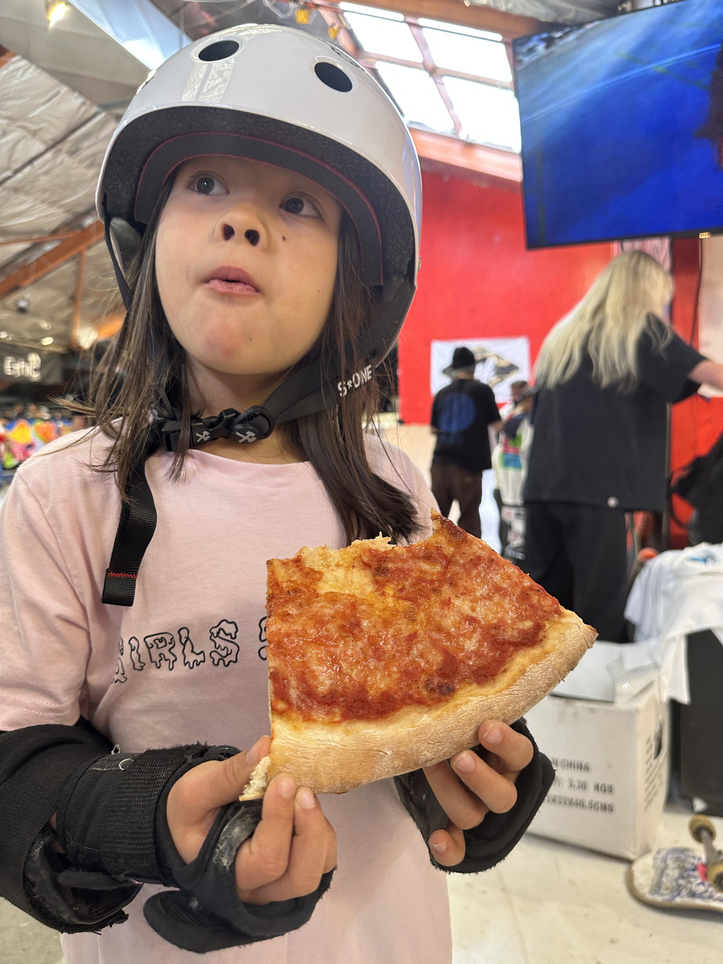 Child wearing a helmet and protective gear holding a slice of pizza indoors.