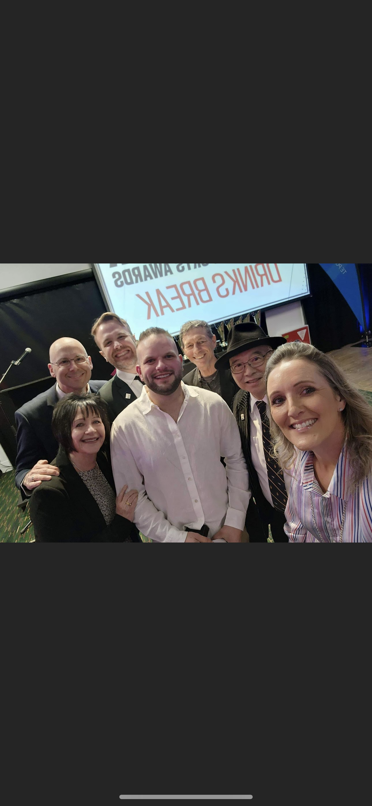 Group of people smiling at a social event with a "Drinks Break" sign in the background.