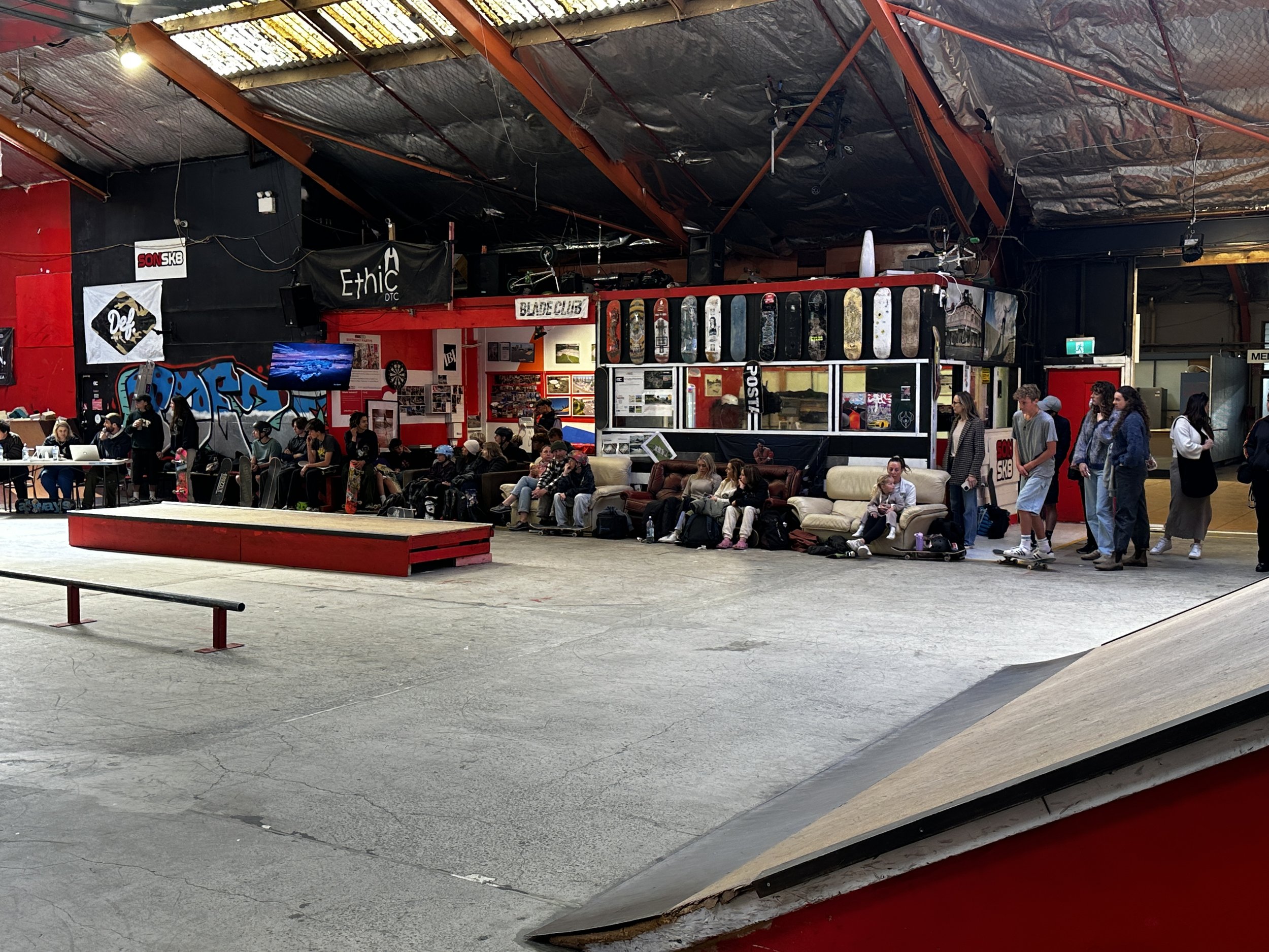 Indoor skate park with ramps and rails, surrounded by people seated and standing. Skateboards and graffiti decorate the walls; a row of skateboards is displayed above them. A television is mounted on the wall, and various posters and banners are visible.