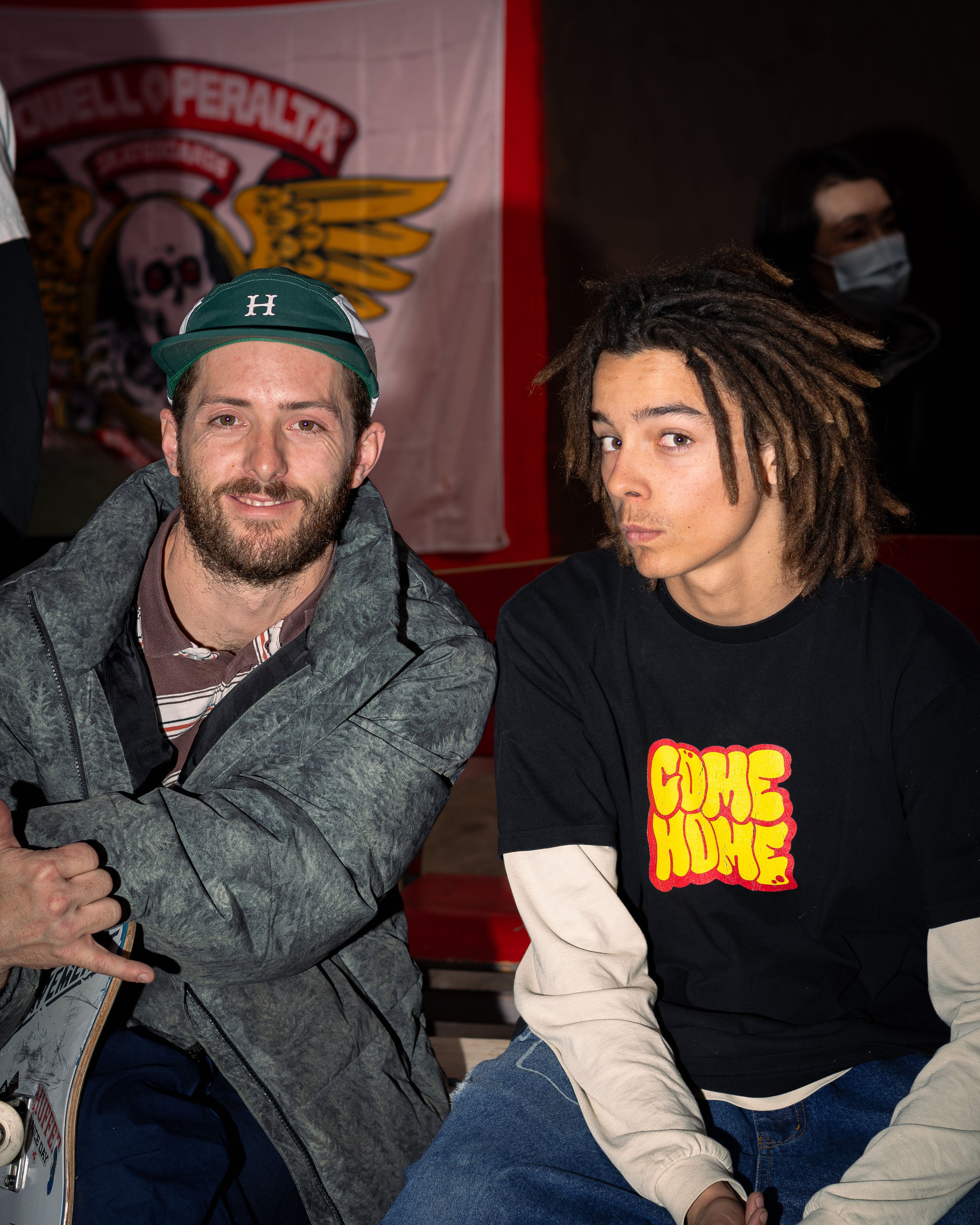 Two men sitting together indoors, one with a skateboard, wearing casual clothing and a cap. A Powell Peralta banner is visible in the background. The man on the left is making a shaka sign with his hand. A person in a mask is partially visible in the