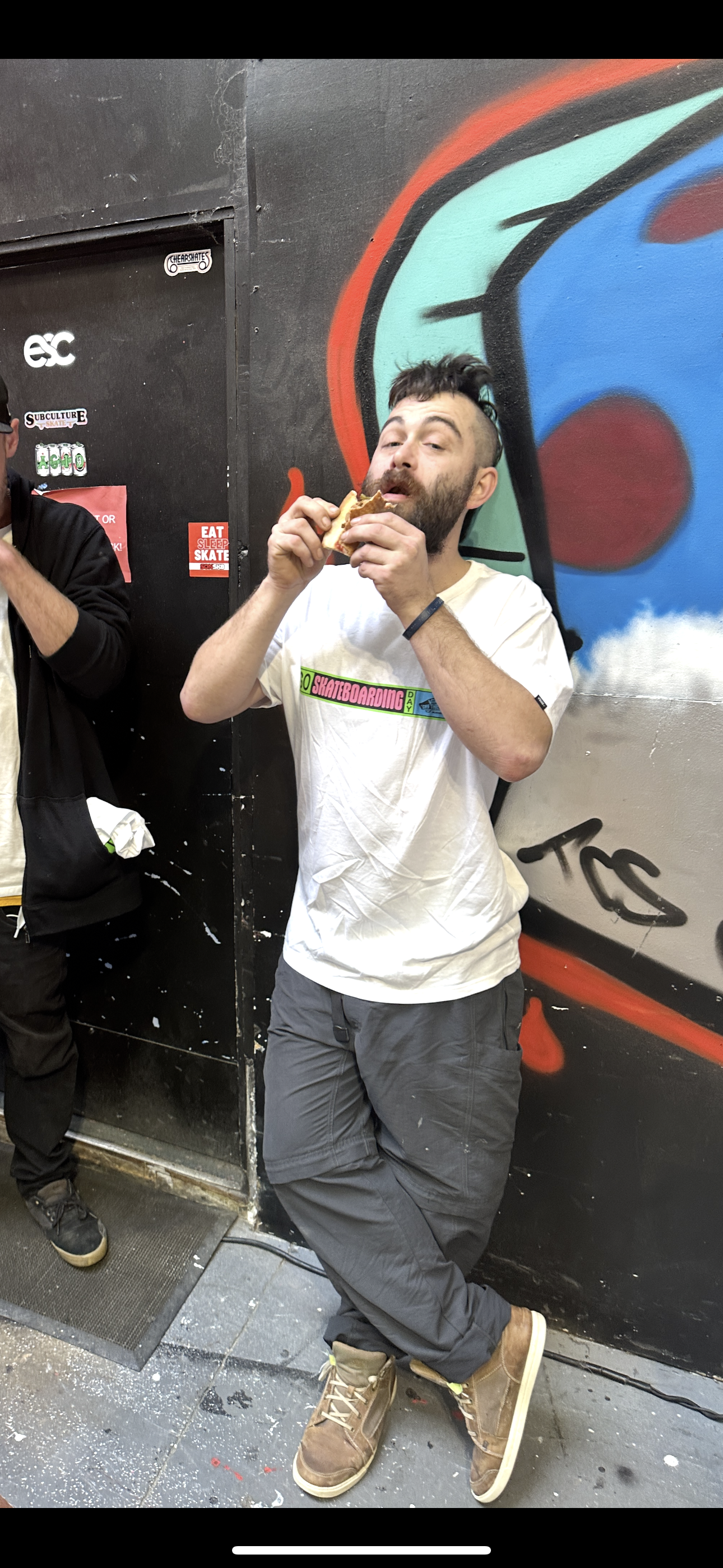 Person eating a sandwich in front of graffiti wall, wearing a white t-shirt and brown shoes.