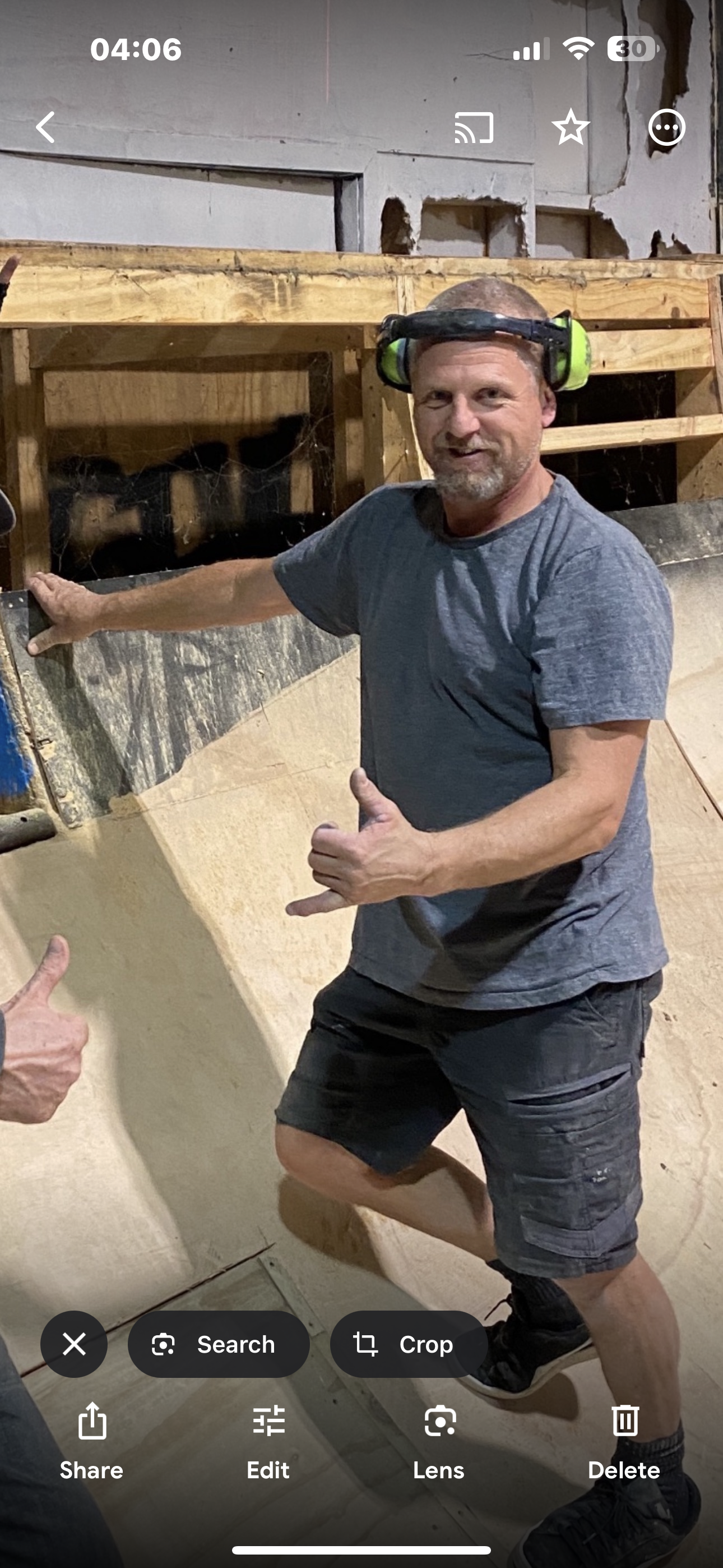 Man wearing ear protection, making a hand gesture, standing indoors near a wooden ramp in a workshop setting.