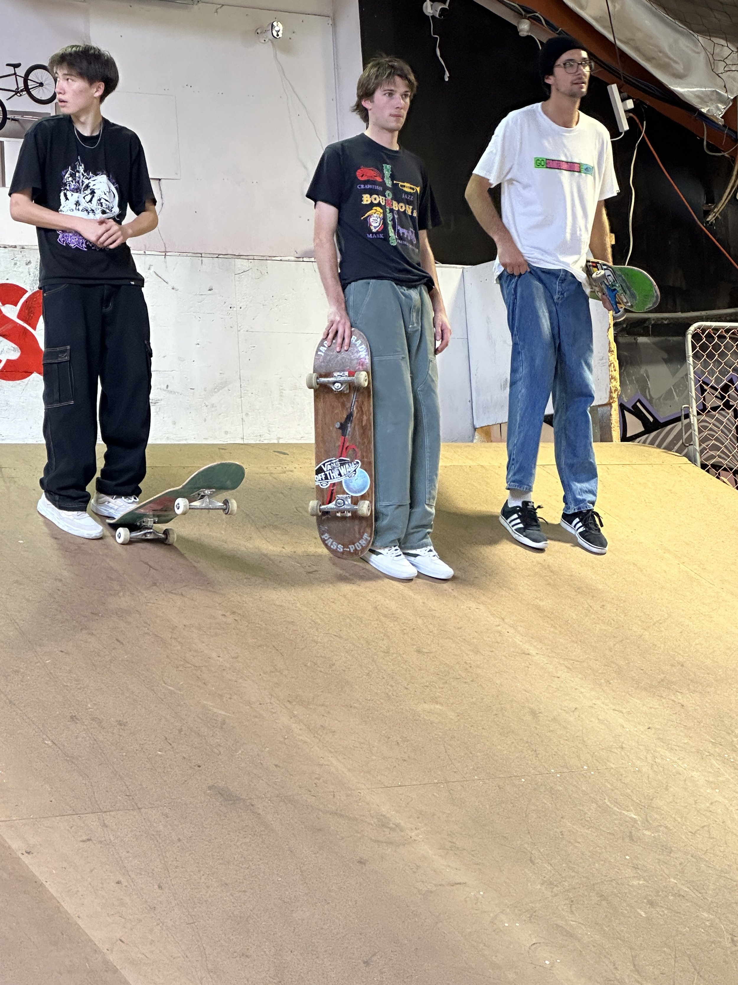 Three skaters standing on an indoor skate ramp, each holding or standing by a skateboard, wearing casual clothing.