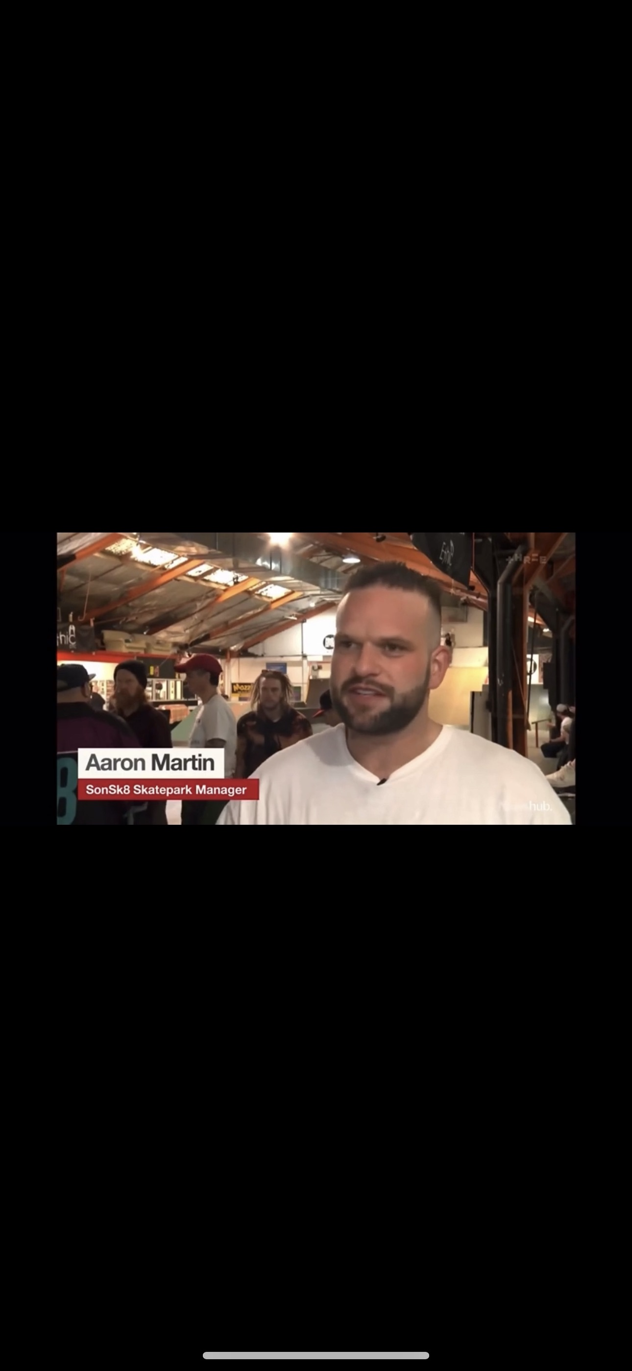 A person in a white shirt stands inside a skatepark with other people visible in the background. The setting includes metal beams and overhead lights. The text overlay reads: "Aaron Martin, SonSk8 Skatepark Manager."
