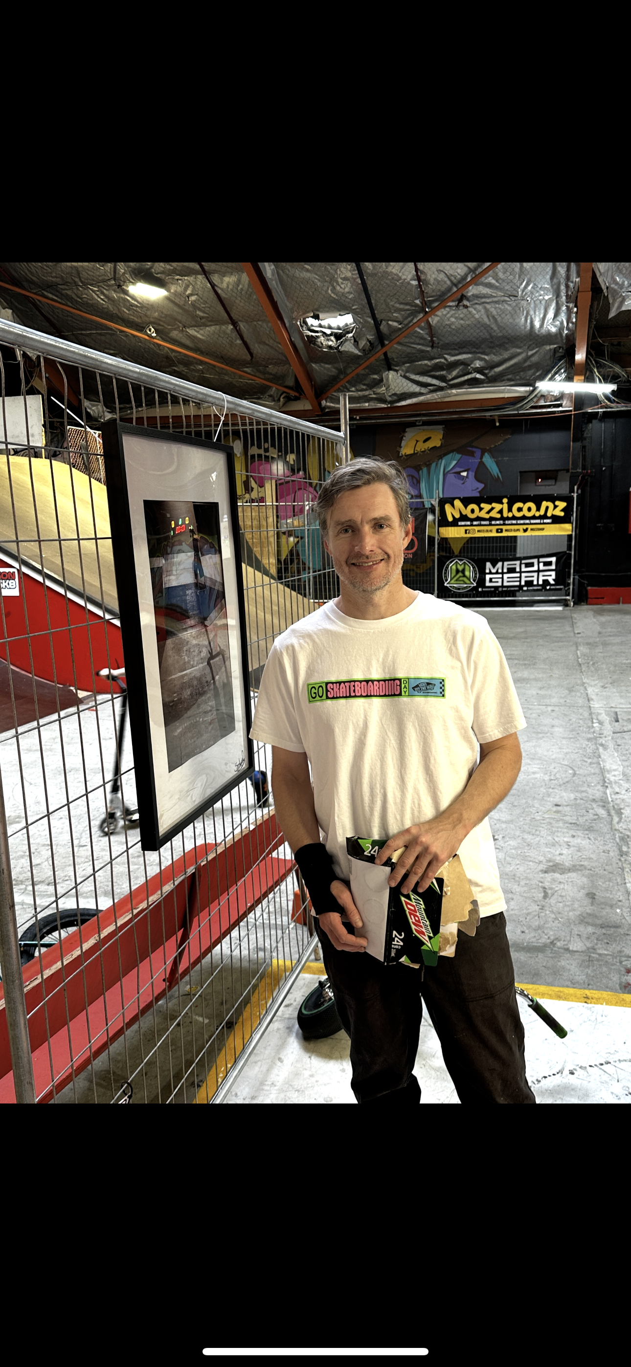 Man holding mountain dew case in indoor skate park, wearing 'Go Skateboarding' t-shirt, standing next to framed photo on fence.