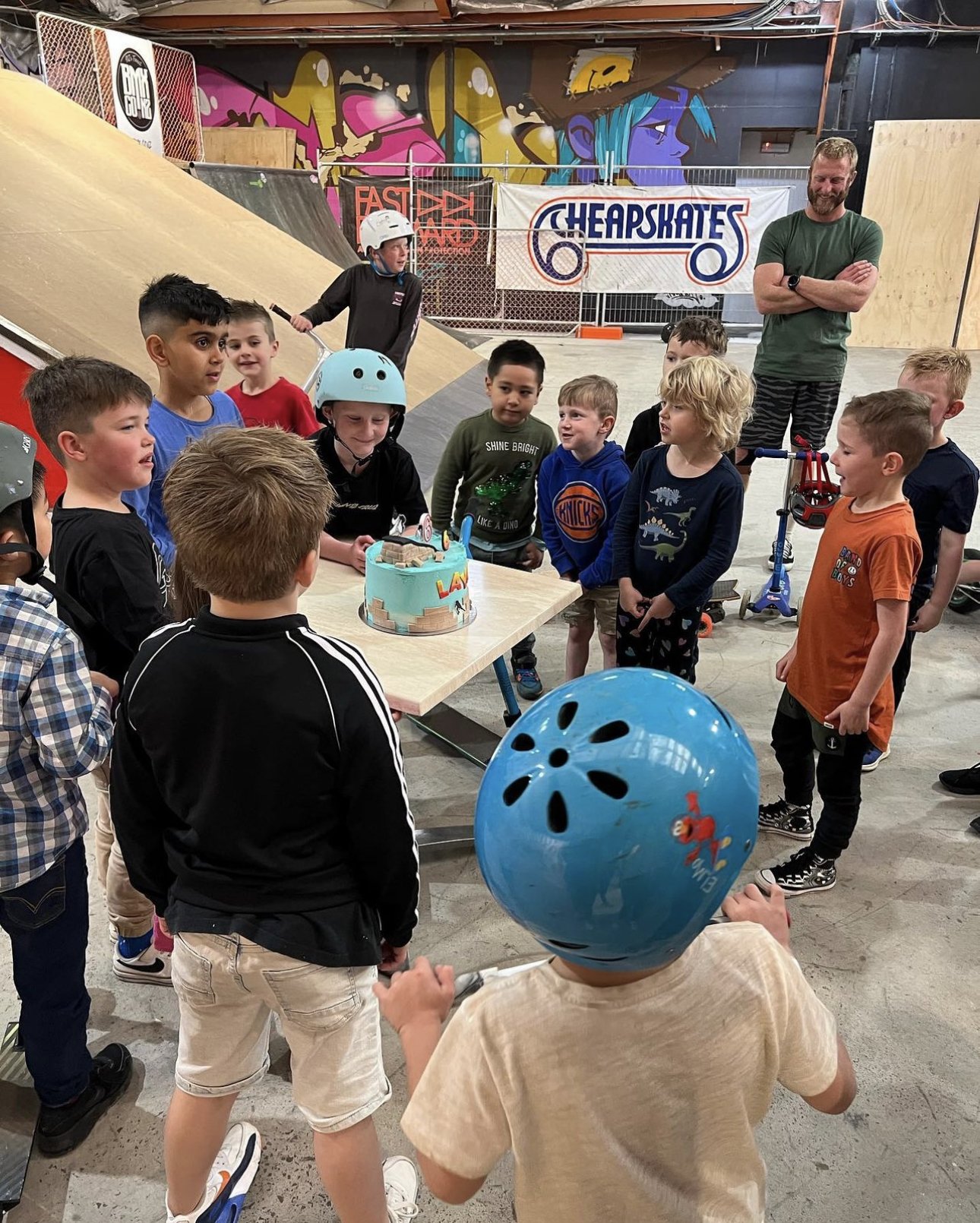 A group of children gather around a birthday cake at an indoor skate park. Some children are wearing helmets, and there are skateboards and scooters visible. An adult supervises in the background.