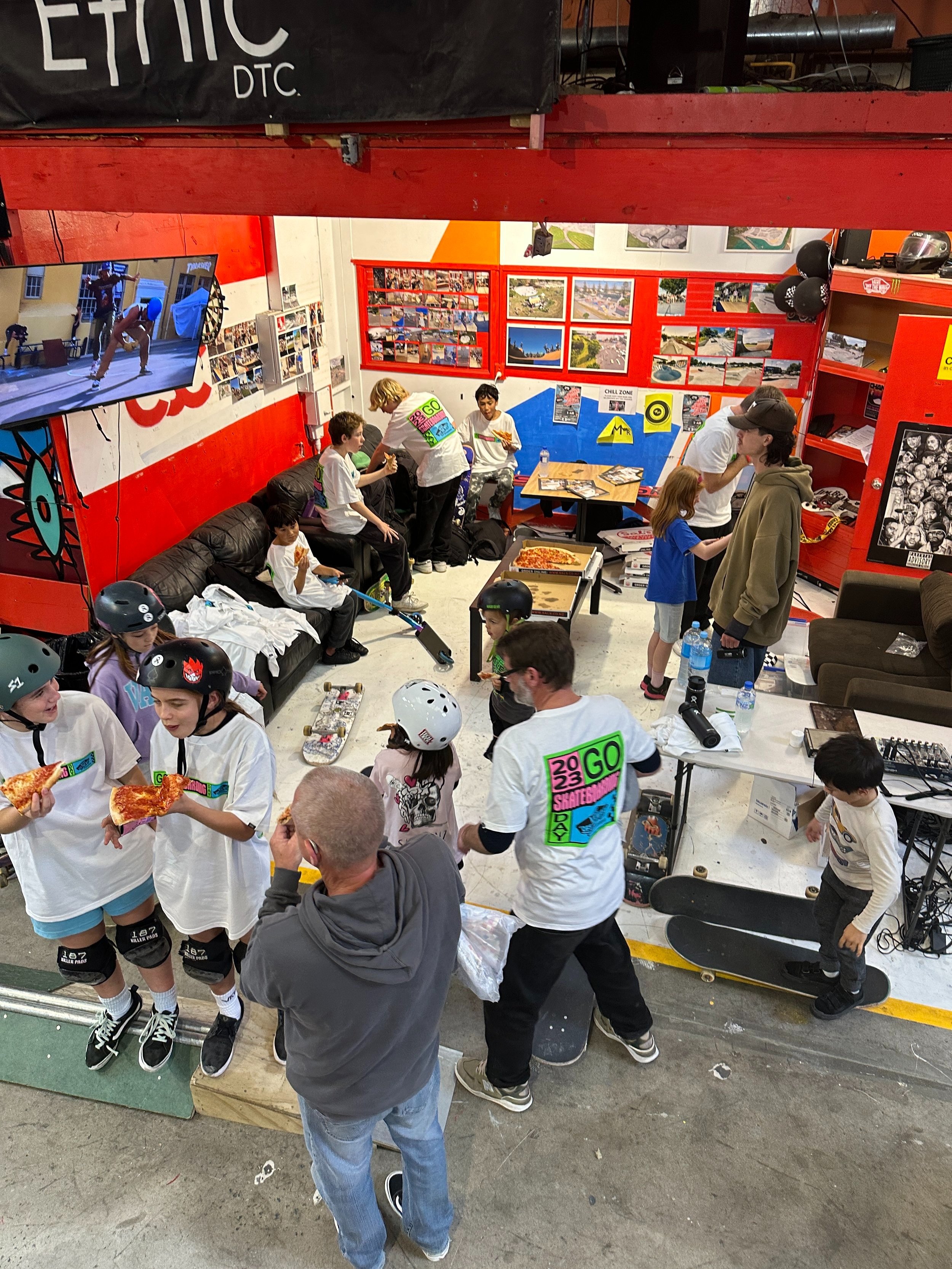 Indoor skateboarding event with children wearing helmets and eating pizza, surrounded by colorful walls and skateboards, featuring posters and photographs.