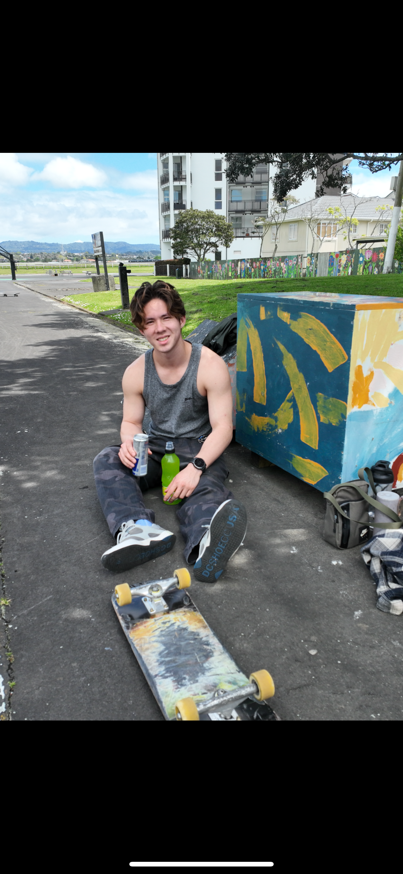 Young man sitting on a sidewalk holding drinks next to a skateboard, with artwork on a box and urban scenery in the background.