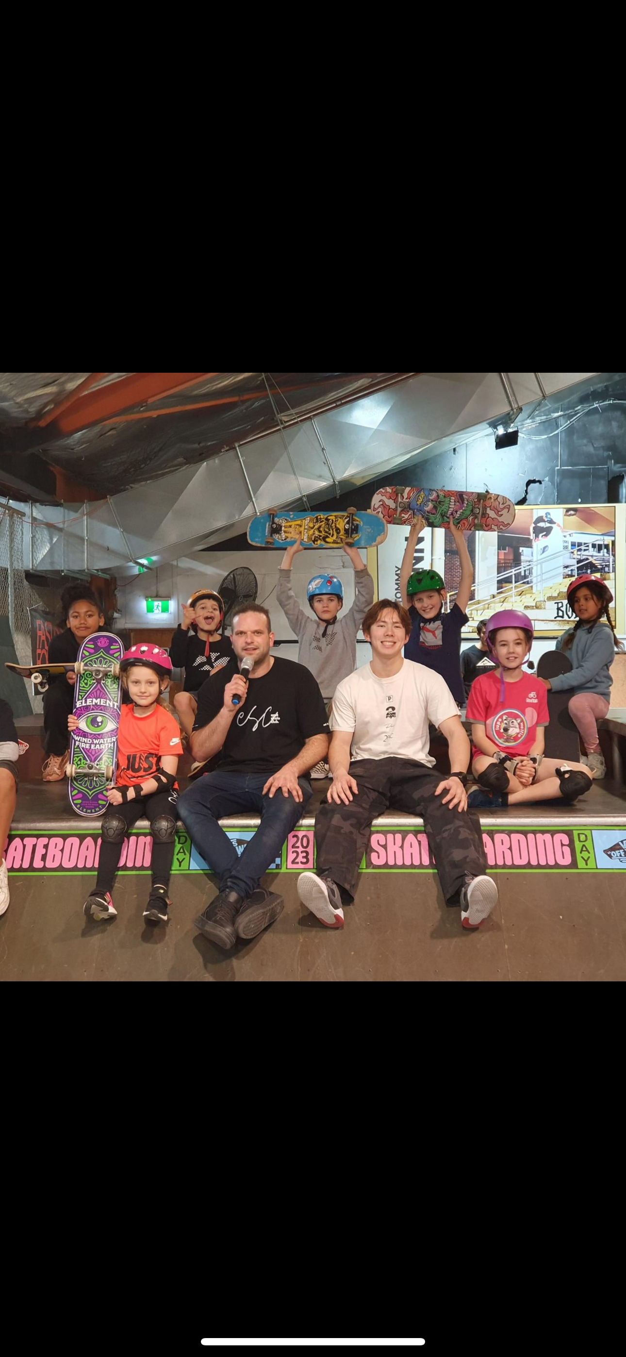 Group of children and adults posing with skateboards indoors, wearing helmets.