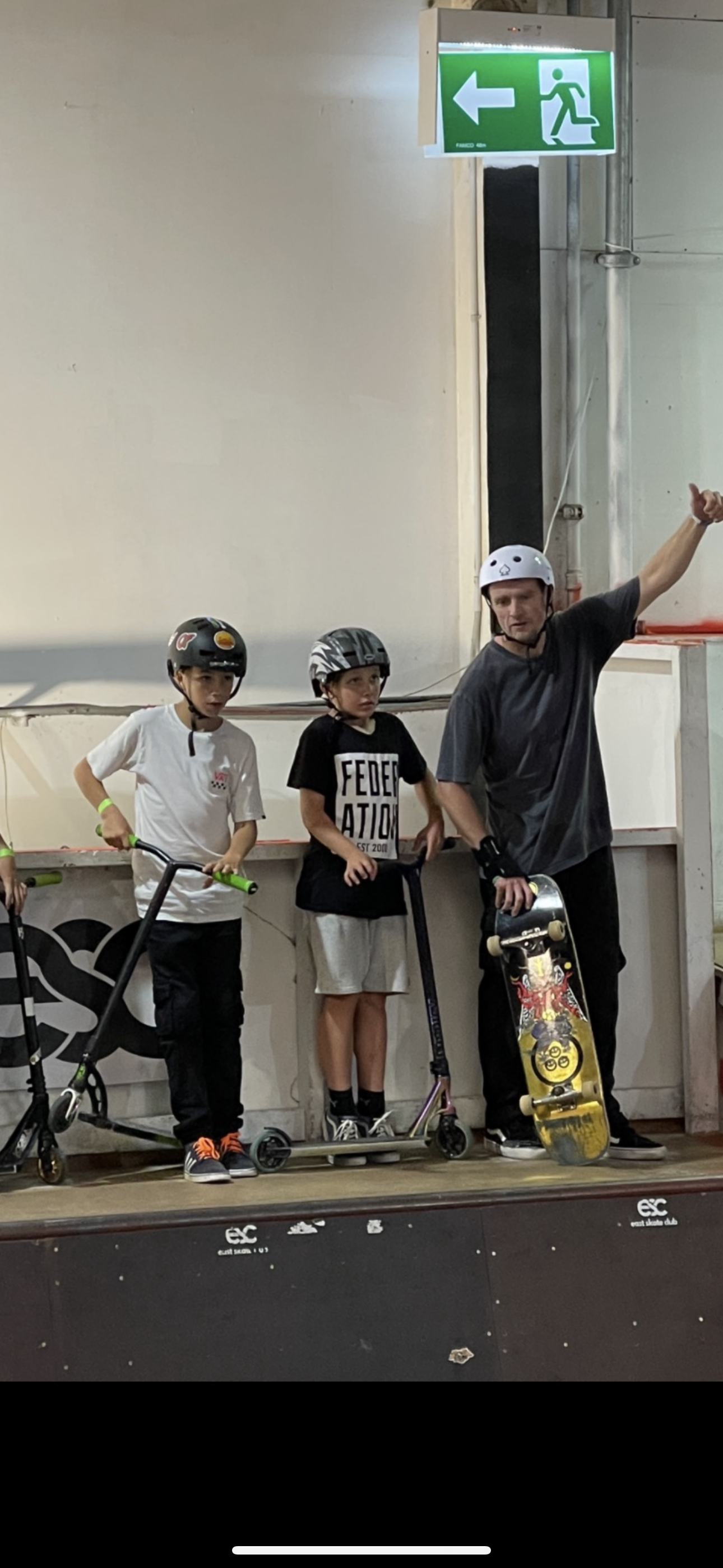 Two children on scooters and an adult with a skateboard standing in a skate park under an exit sign.