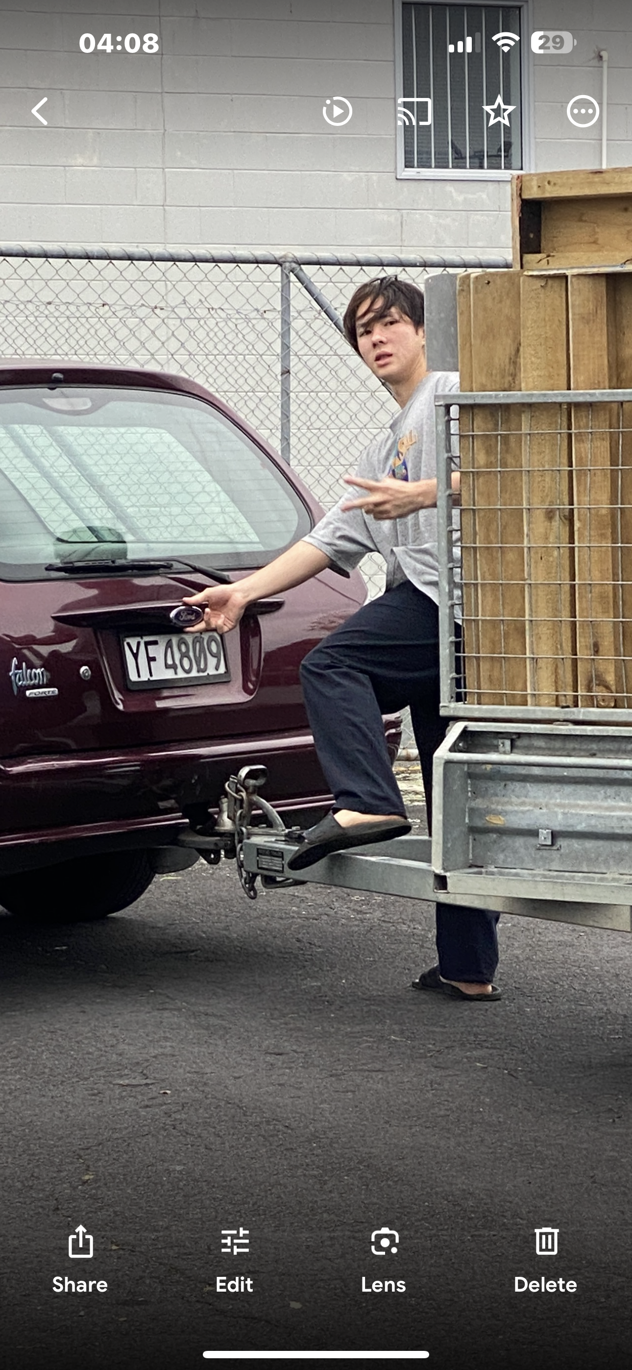 Person standing near the back of a red Ford car with a trailer attached, holding the Ford emblem, and making a gesture. A fence and building in the background.