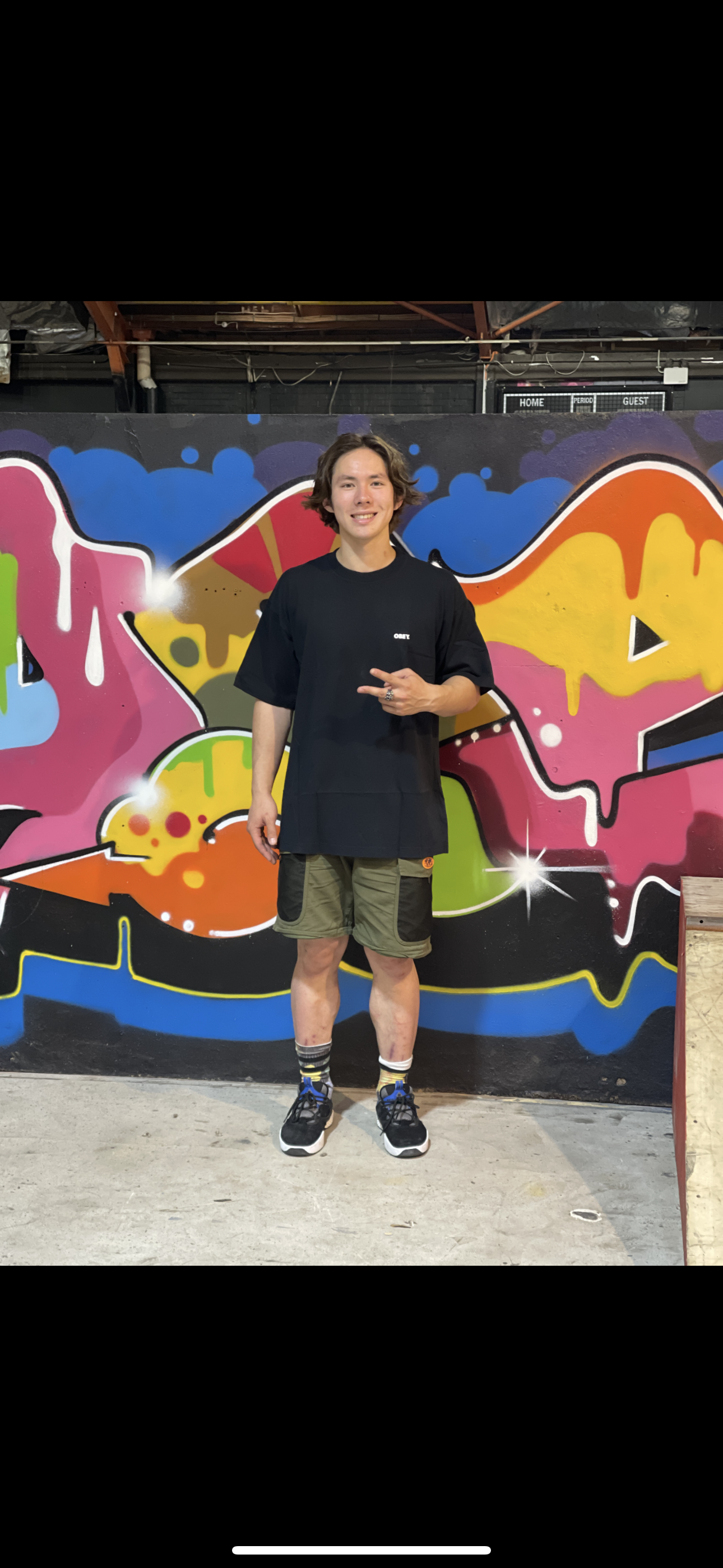 Person standing in front of colorful graffiti wall, wearing a black t-shirt, green shorts, and sneakers, with peace sign gesture.