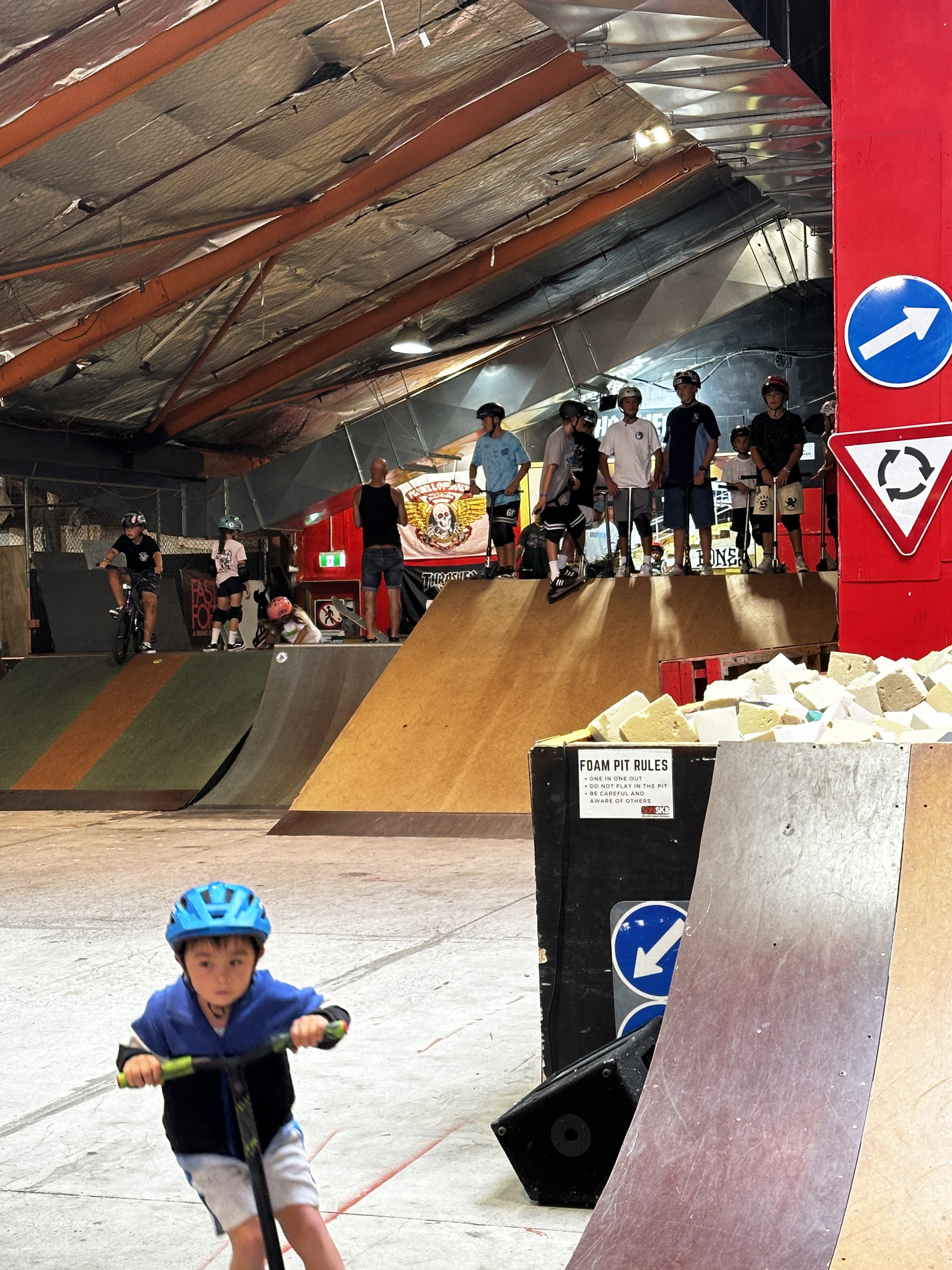 Indoor skate park with ramps, a young boy on a scooter wearing a helmet, and a group of people standing on a ramp in the background. Foam pit with signs visible.