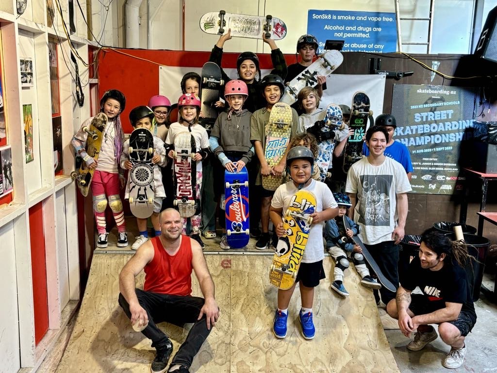 Group of skateboarders posing indoors with boards, wearing helmets and protective gear, in front of signs for a street skateboarding championship.