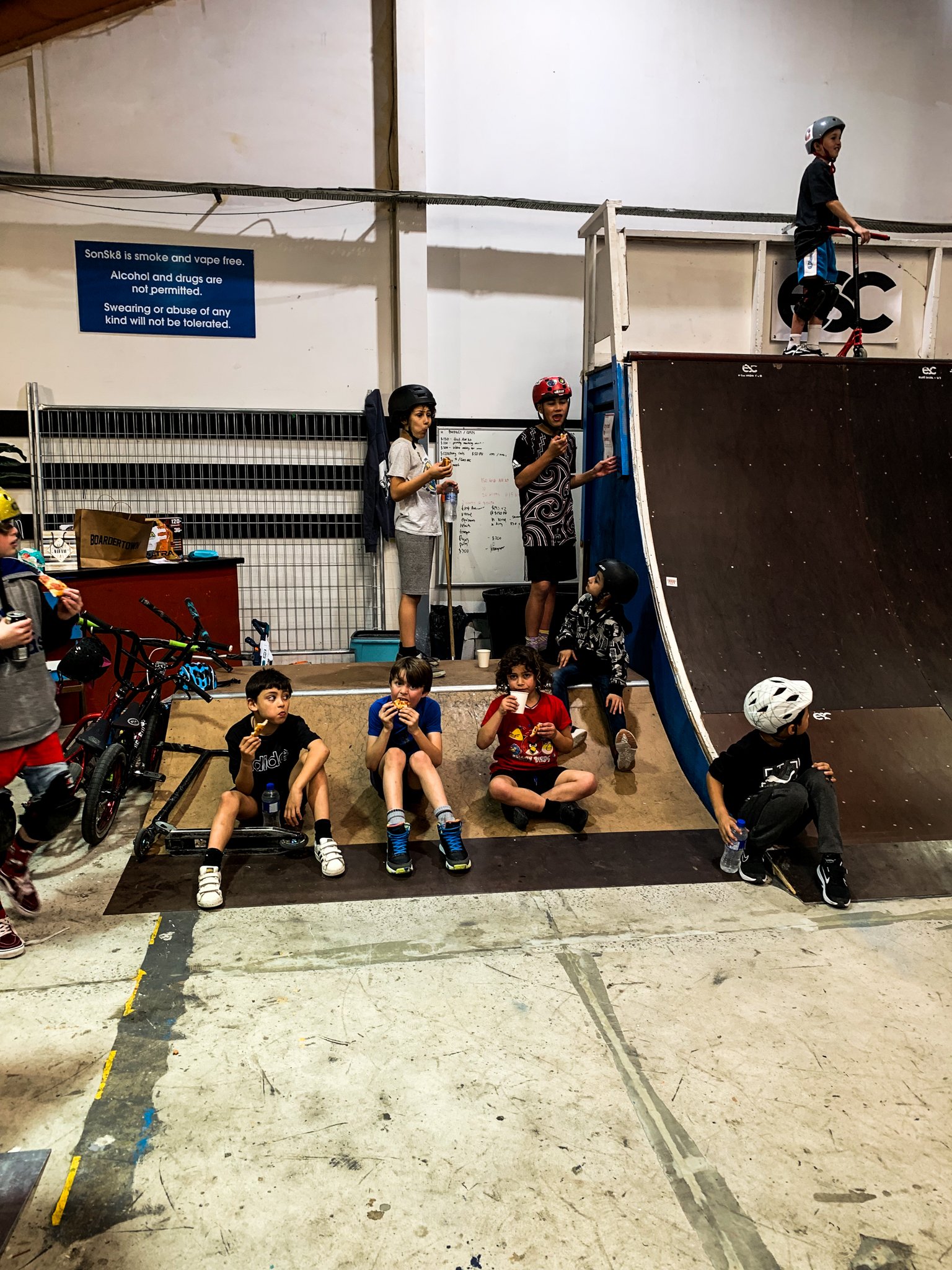 Children wearing helmets at an indoor skate park, some holding snacks and sitting near a ramp, with scooters and bikes nearby, under a sign prohibiting smoking and drugs.