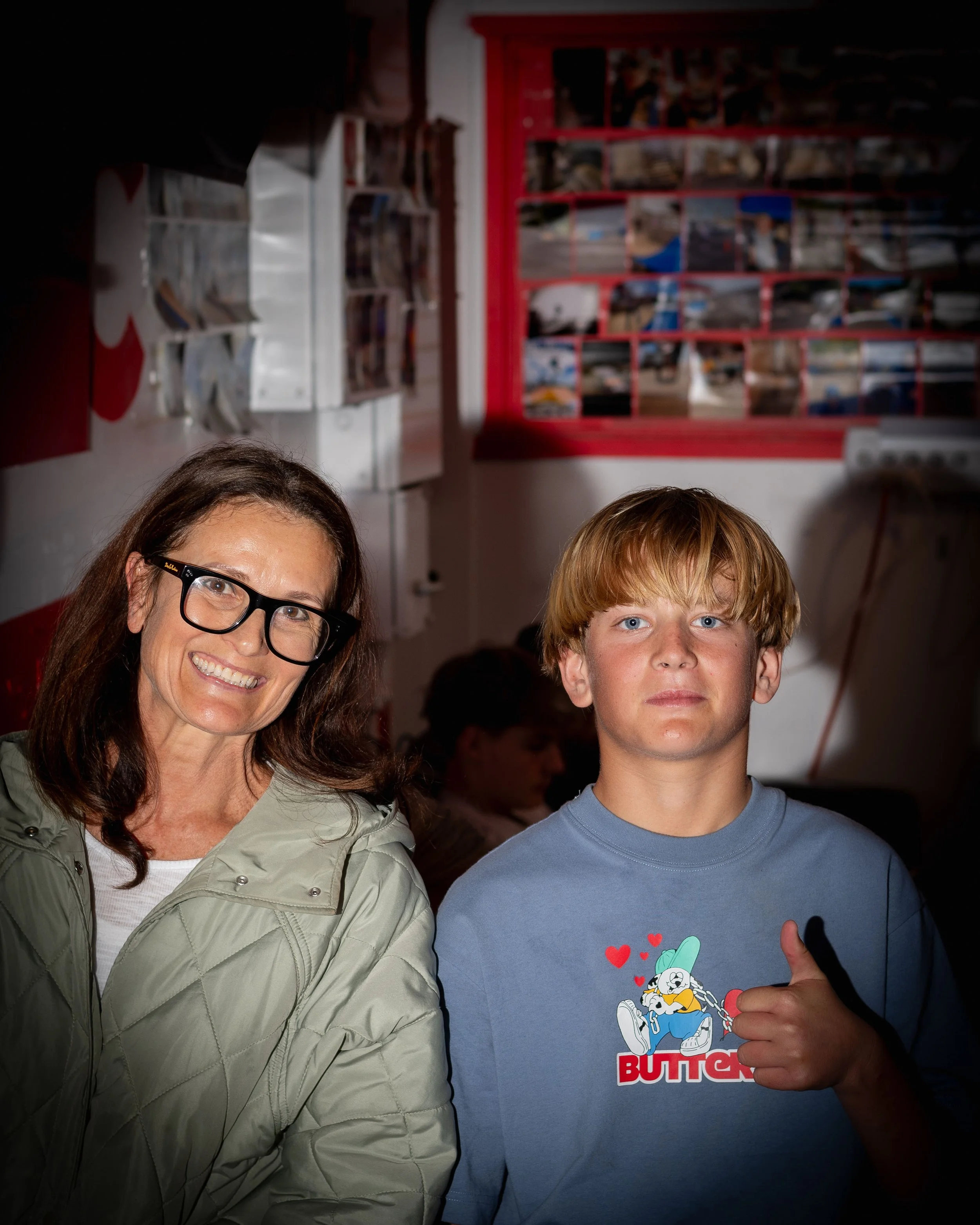 A woman and a young boy standing indoors. The woman is smiling and wearing glasses and a light green jacket. The boy has a straight face, giving a thumbs up, and is wearing a blue t-shirt with a cartoon design and the word "BUTTON." Behind them, ther