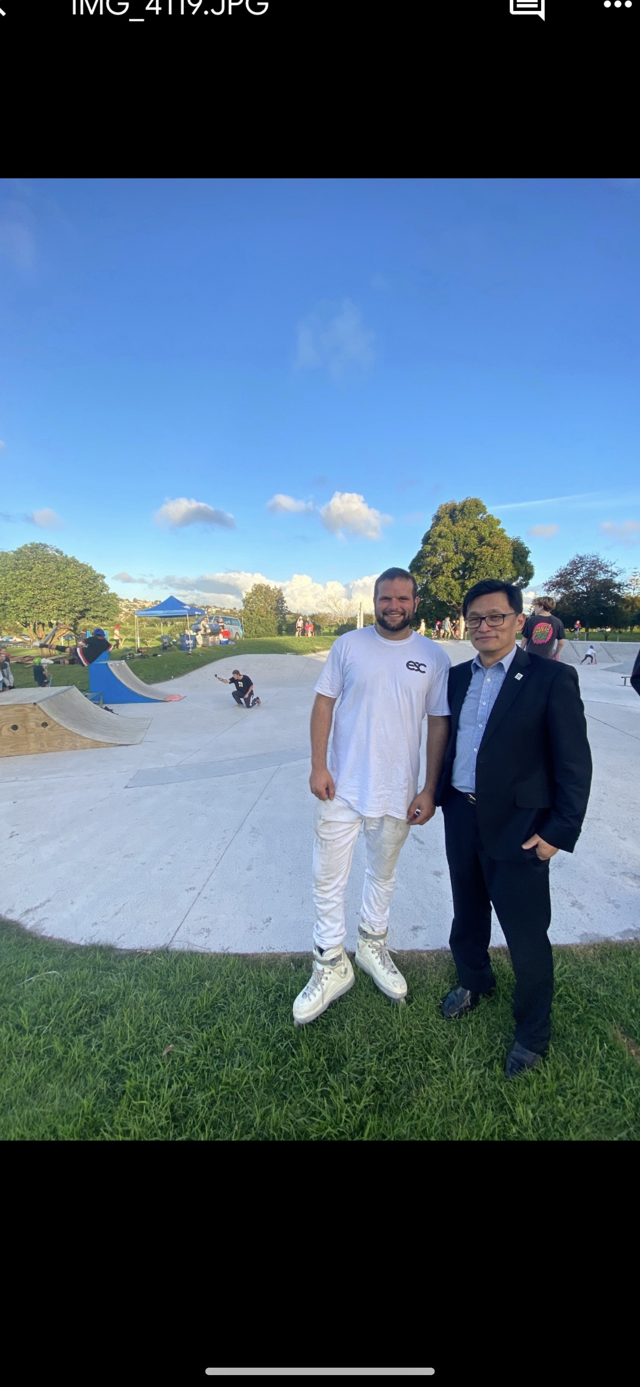 Two men standing together at a skate park; one is dressed casually in a white shirt and pants, the other in a suit. The background shows people skating, trees, and a blue sky.