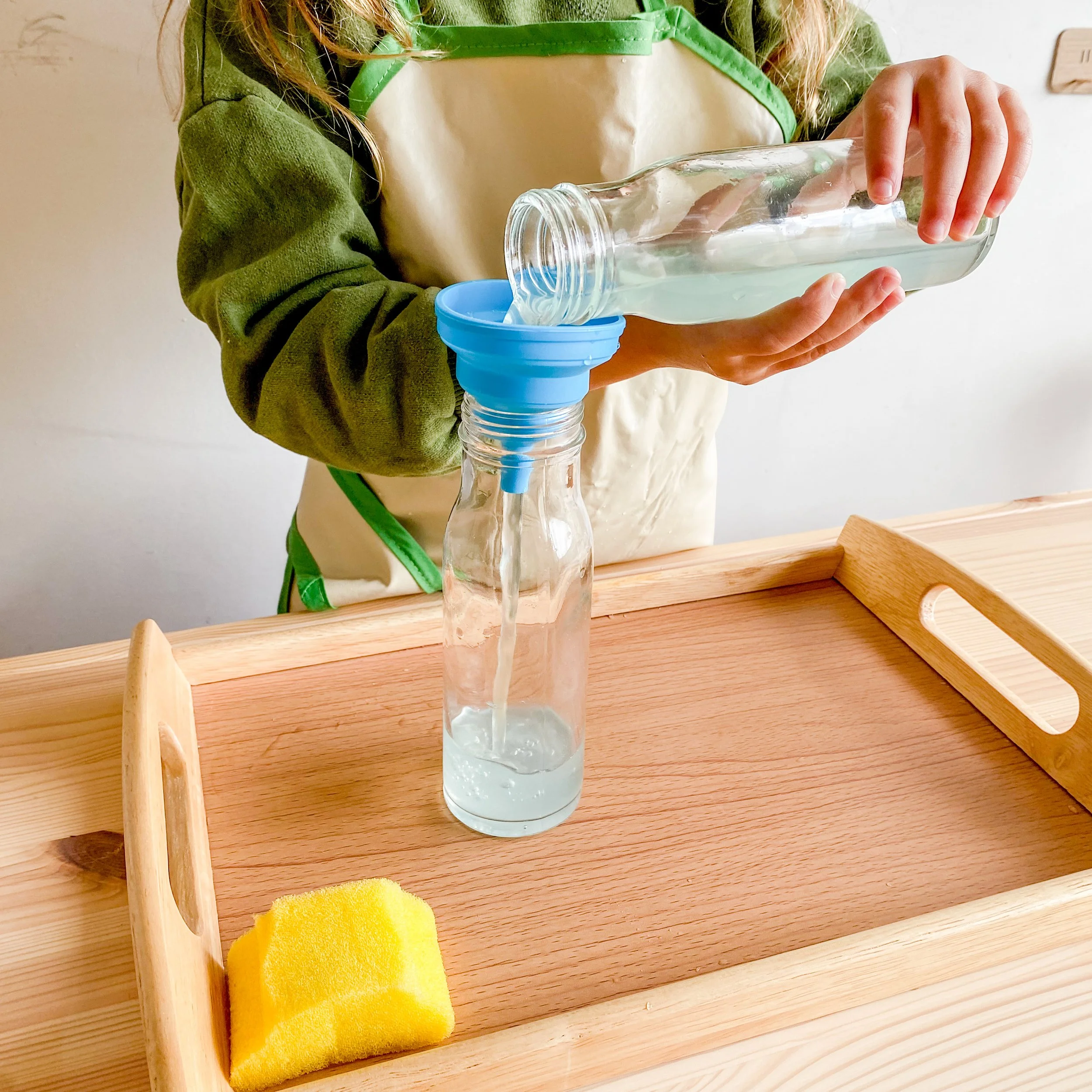 A child filling an empty glass bottle with water from a larger bottle using a blue silicone funnel, on a wooden table with a yellow sponge nearby, illustrating the Pouring Activity in the Practical Life Montessori Curriculum.