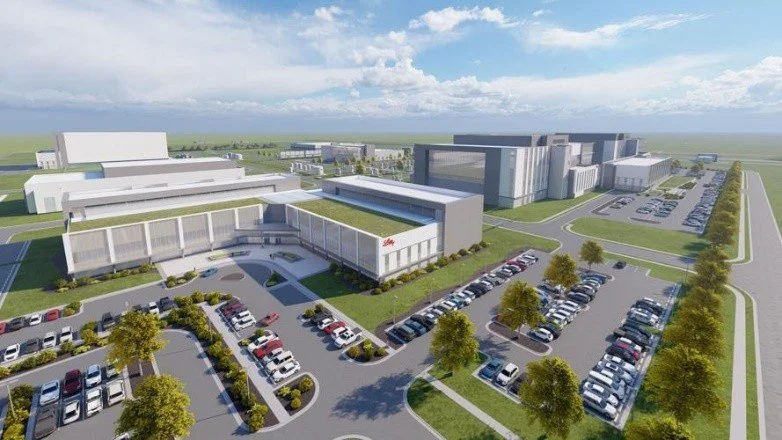 Aerial view of a modern industrial complex with multiple large buildings and adjacent parking lots, surrounded by green lawns. The sky is partly cloudy, indicating a sunny day.