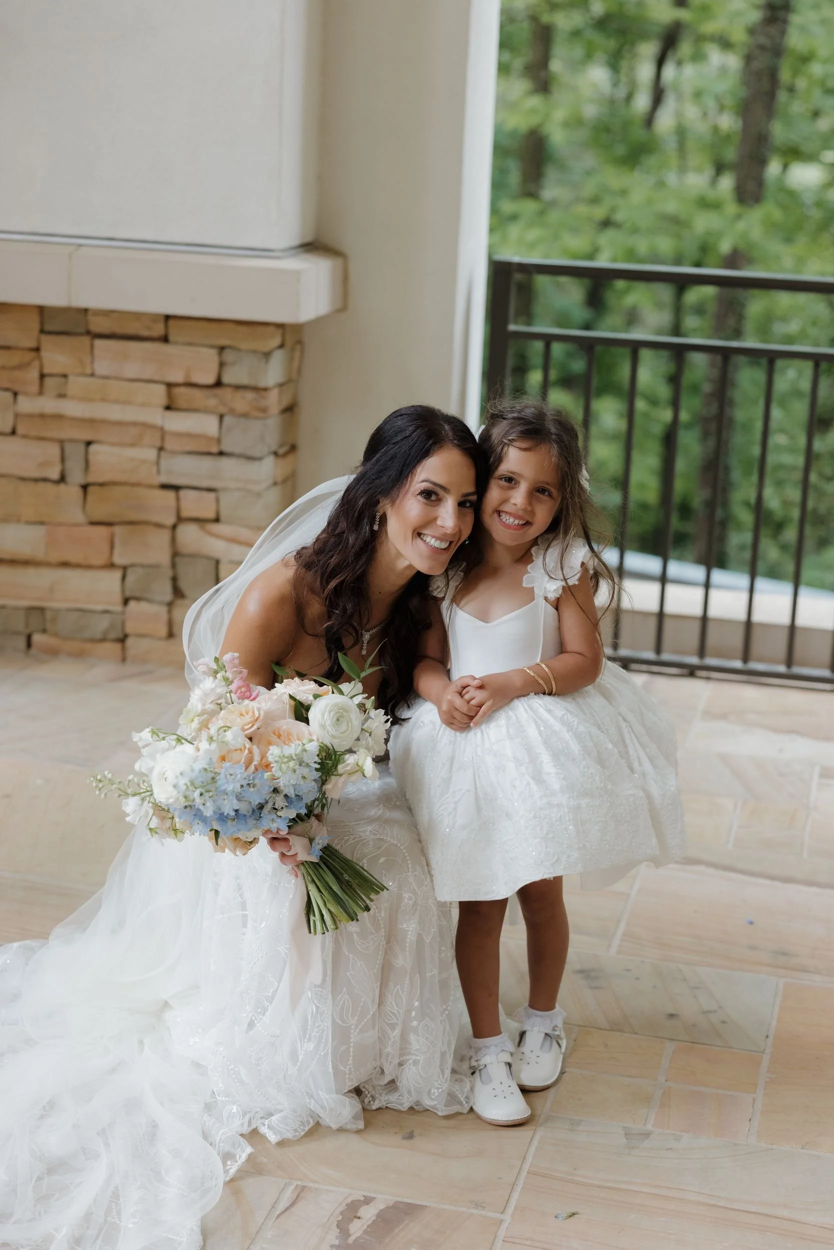 Bride kneeling beside a flower girl on a stone patio, holding a bouquet of ivory, blush, and blue blooms