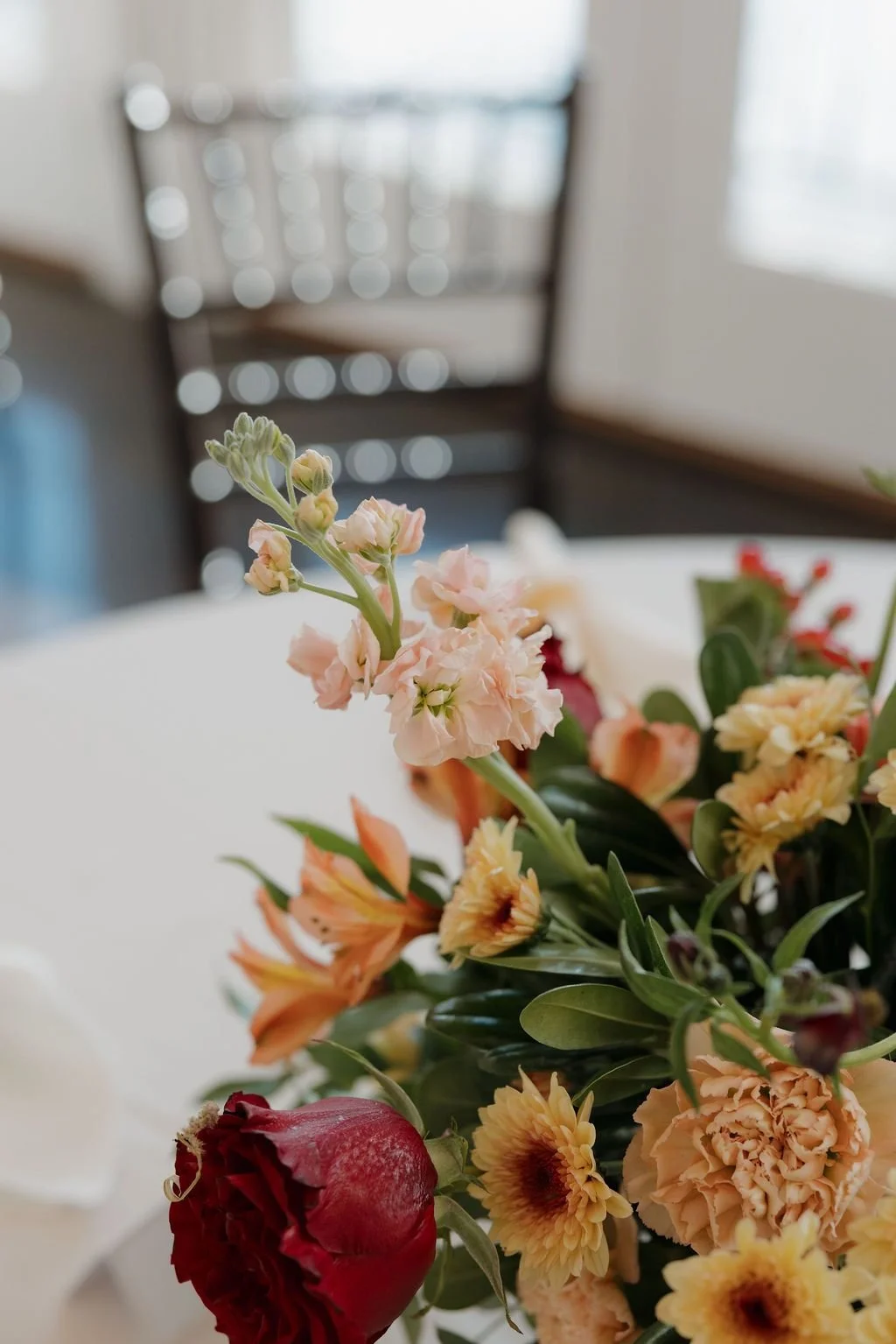 Low centerpiece of peach stock, yellow dahlias, red roses, and greenery on a reception table