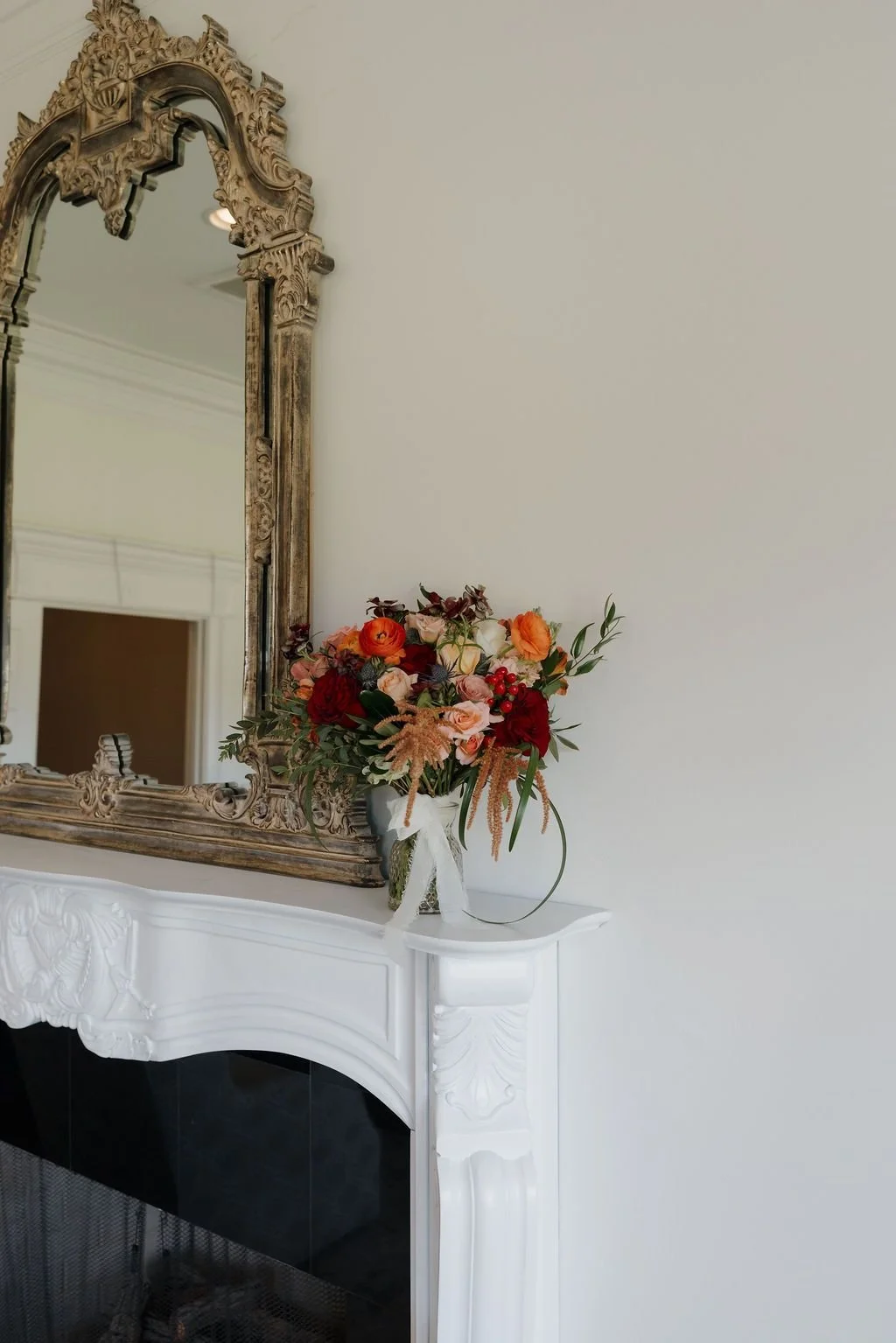 Floral arrangement in a white vase on an ornate fireplace mantel beneath a gilded mirror