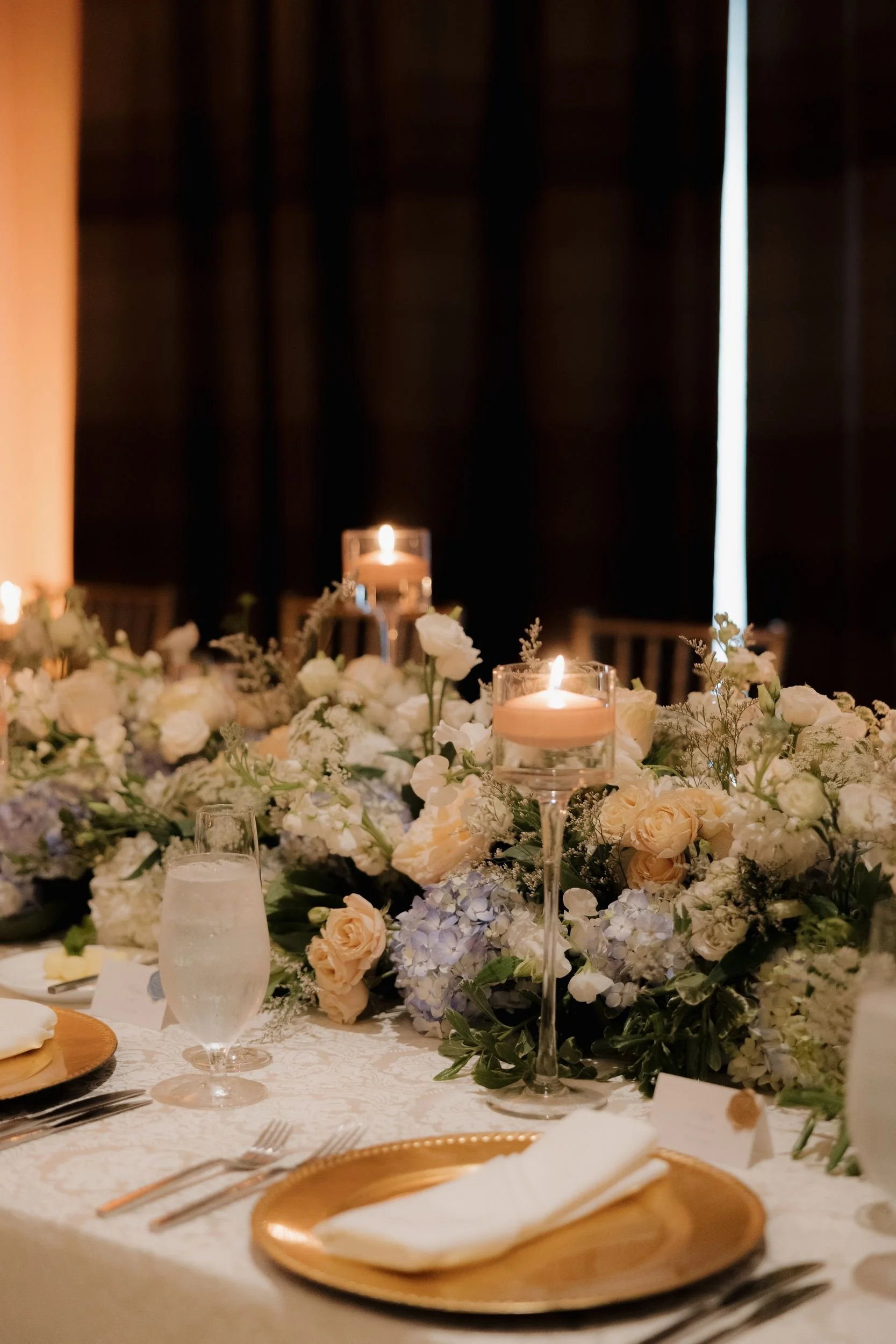 Long reception table with a low floral runner of blue hydrangea, white, and blush blooms, pillar candles, and gold chargers