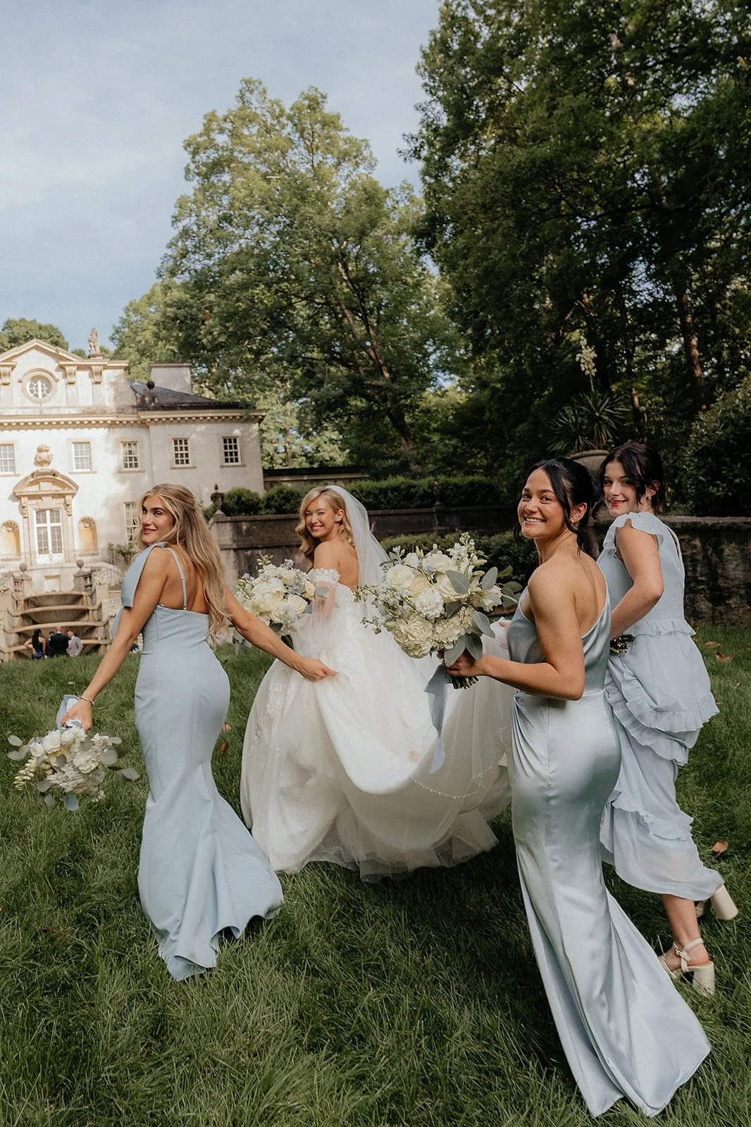 Bride and bridesmaids in light blue dresses walking through a lush garden at Swan House Atlanta, holding white and greenery bouquets, with the historic estate in the background creating a romantic outdoor wedding setting.