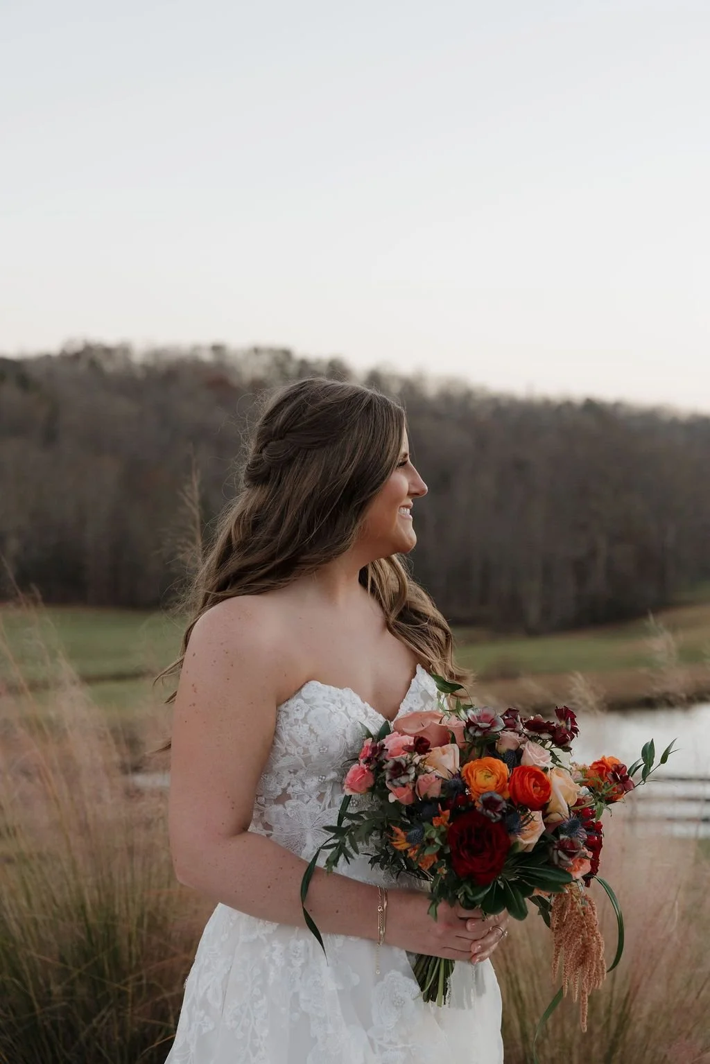 Bride in a lace gown holding a cascading terracotta and peach bouquet in a field by the lake