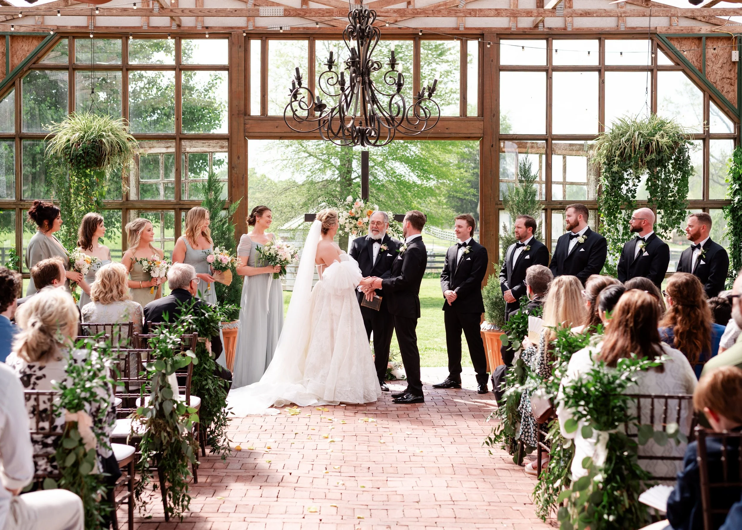 Couple exchanging vows in a greenhouse wedding ceremony with lush greenery, brick aisle, and elegant floral designs.