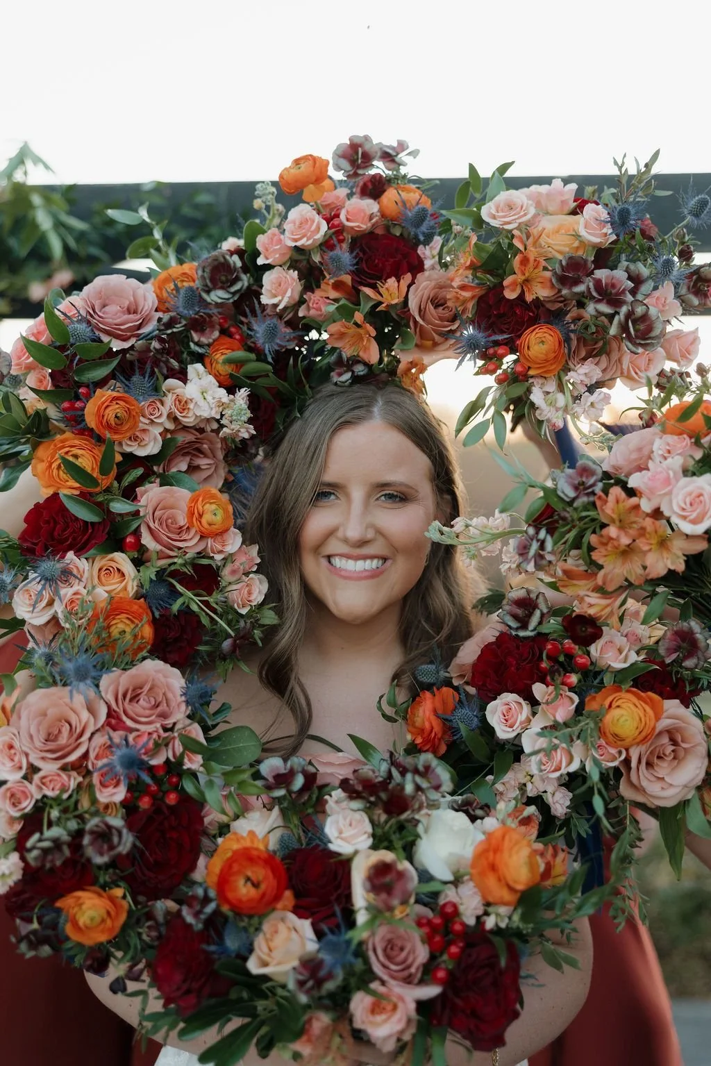 Person framed by a large circular floral hoop densely covered in red, orange, and peach blooms