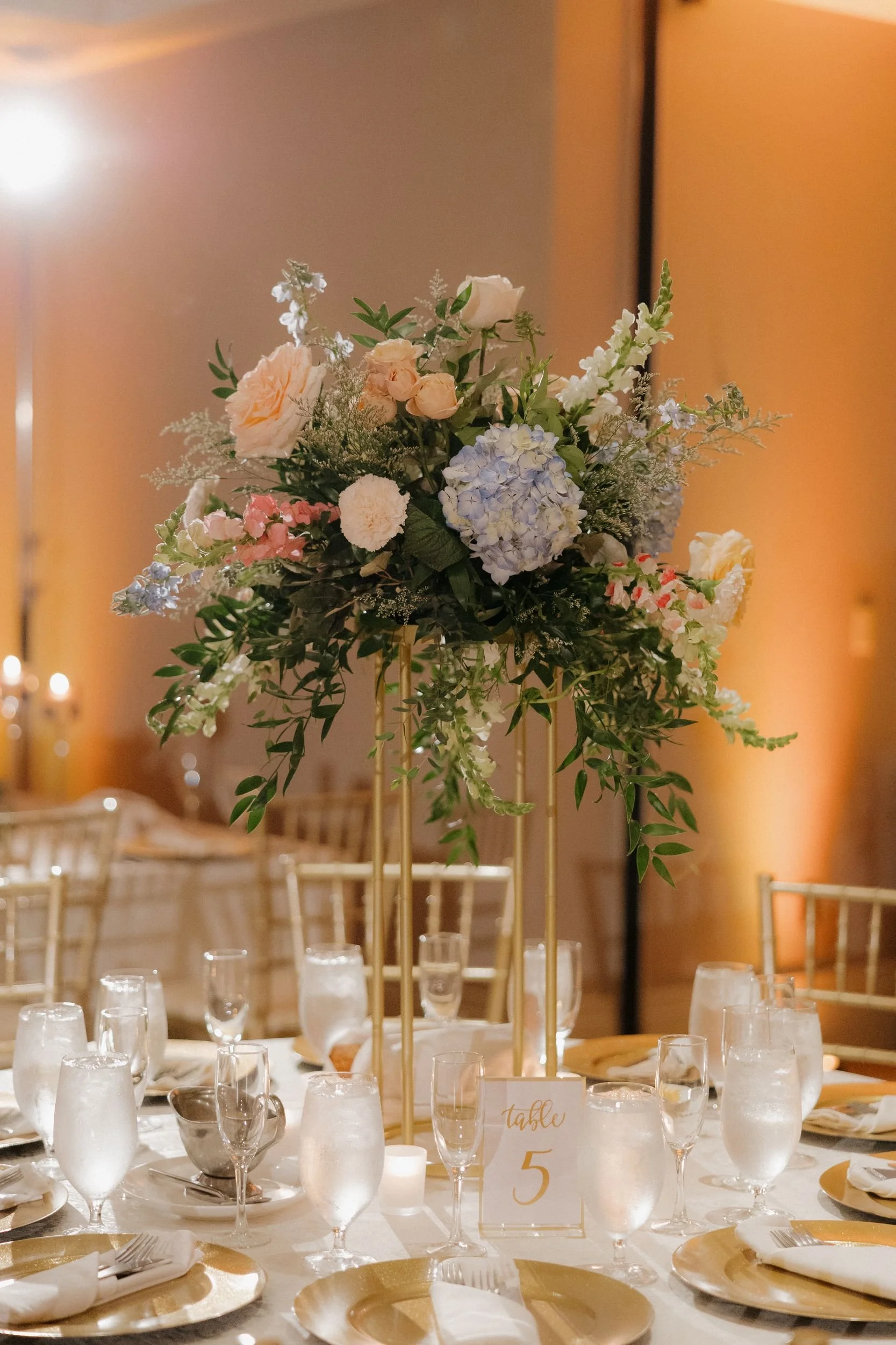 Tall reception centerpiece of peach roses and blue hydrangea on a gold stand above a round table set with gold chargers and glassware