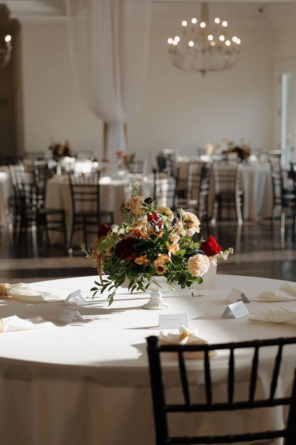 Reception ballroom with round tables, a low peach and burgundy centerpiece, place settings, and a chandelier