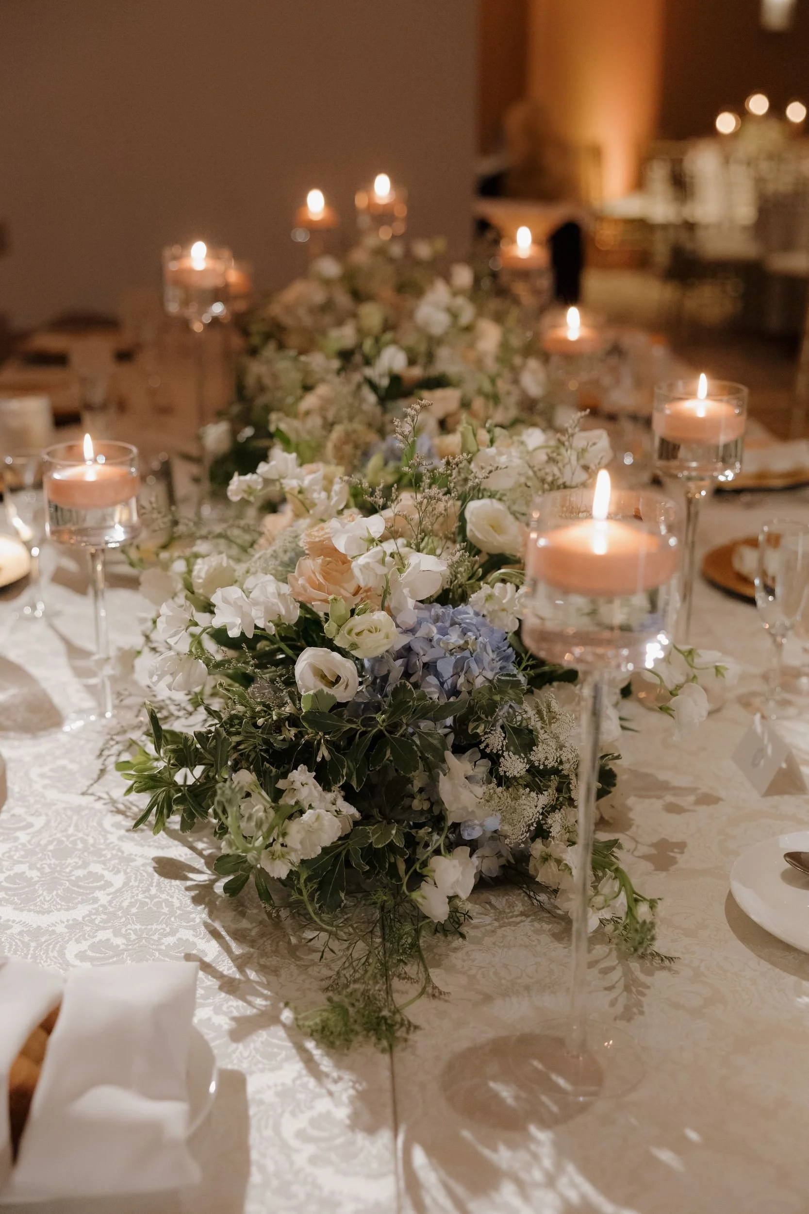 Long banquet table with a low floral garland of white roses and blue hydrangea among floating candles