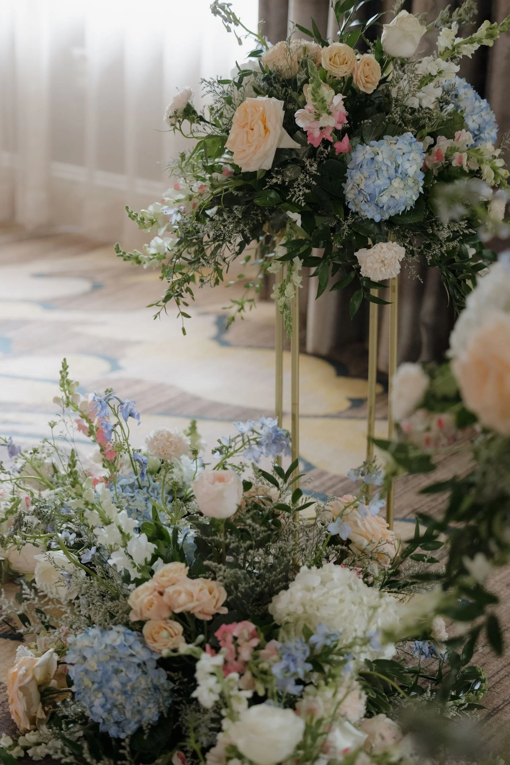 Tall ceremony arrangement on a gold stand with peach roses and blue hydrangea above a lush ground installation of hydrangea and white blooms