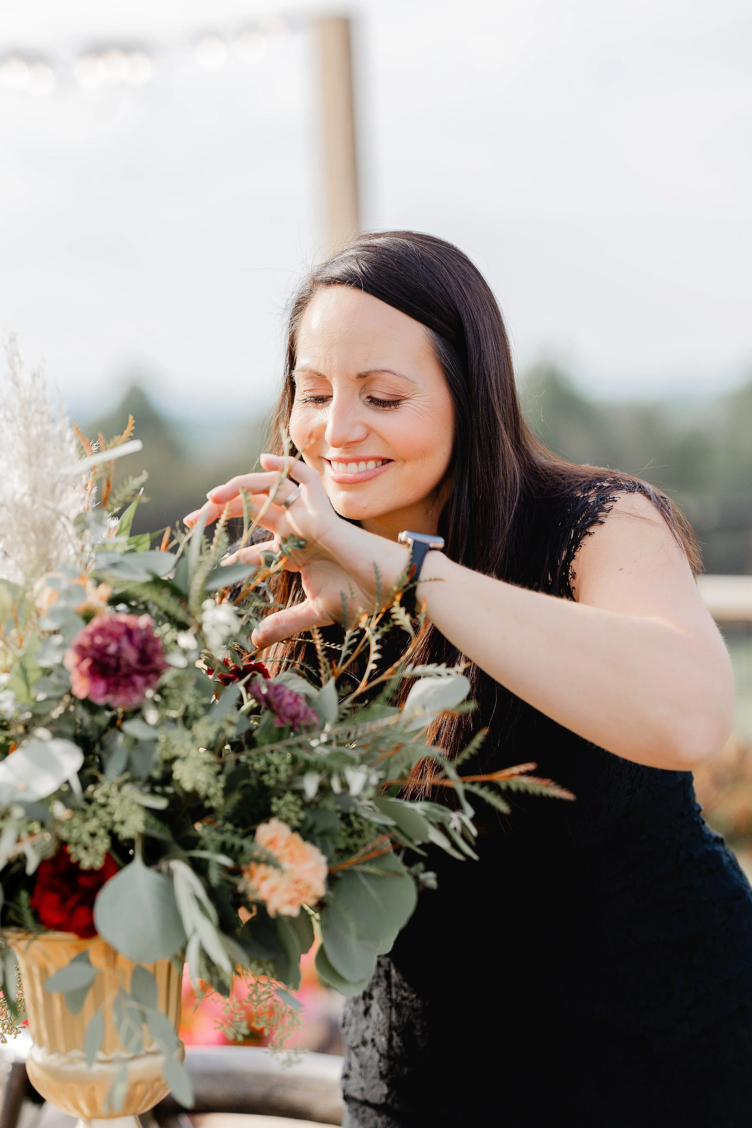 Wedding florist arranging a lush floral centerpiece with greenery, peach, and burgundy blooms, smiling while styling details for an elegant event.