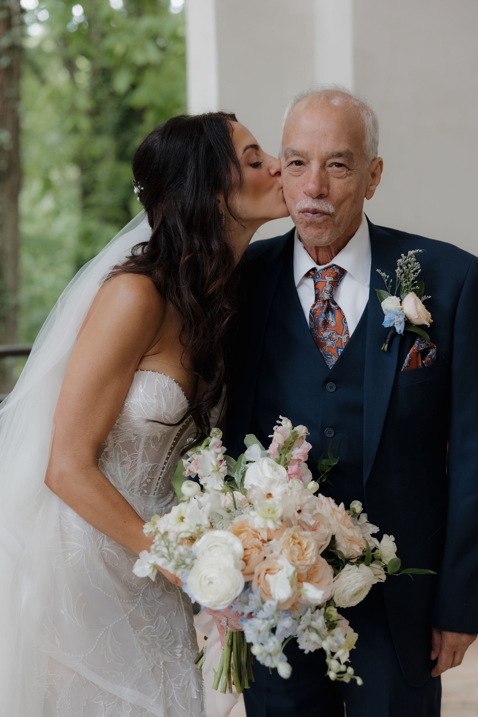 Bride kissing her father's cheek while holding a lush peach and ivory bouquet, father in a navy suit with a boutonniere